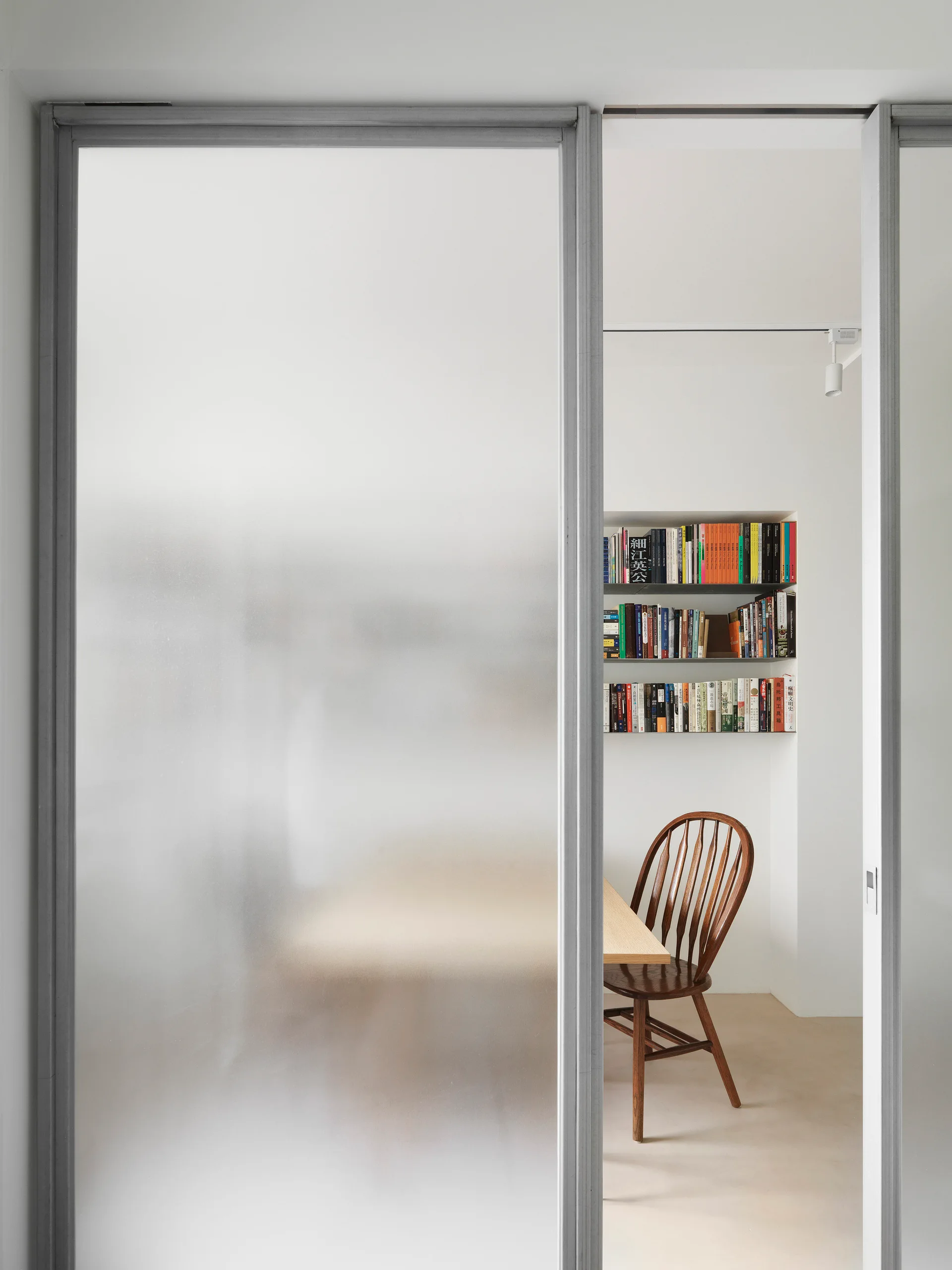 A frosted glass sliding door reveals a minimalist home office featuring a wooden spindle chair and books on a recessed wall shelf.