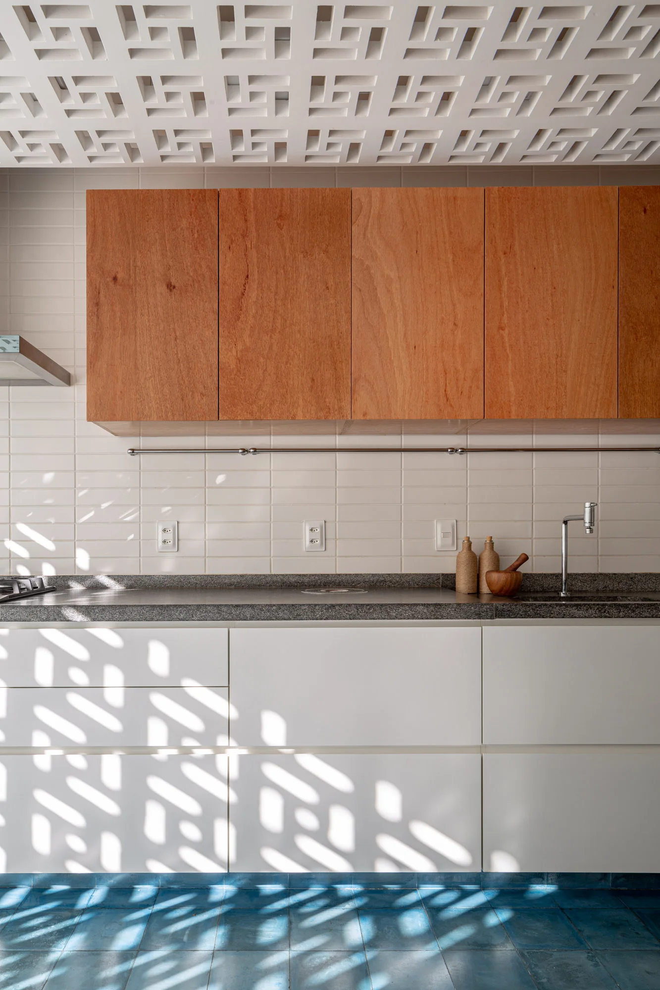 A modern kitchen featuring natural wood upper cabinets, white base cabinets, a grey countertop, and a decorative white geometric screen ceiling.