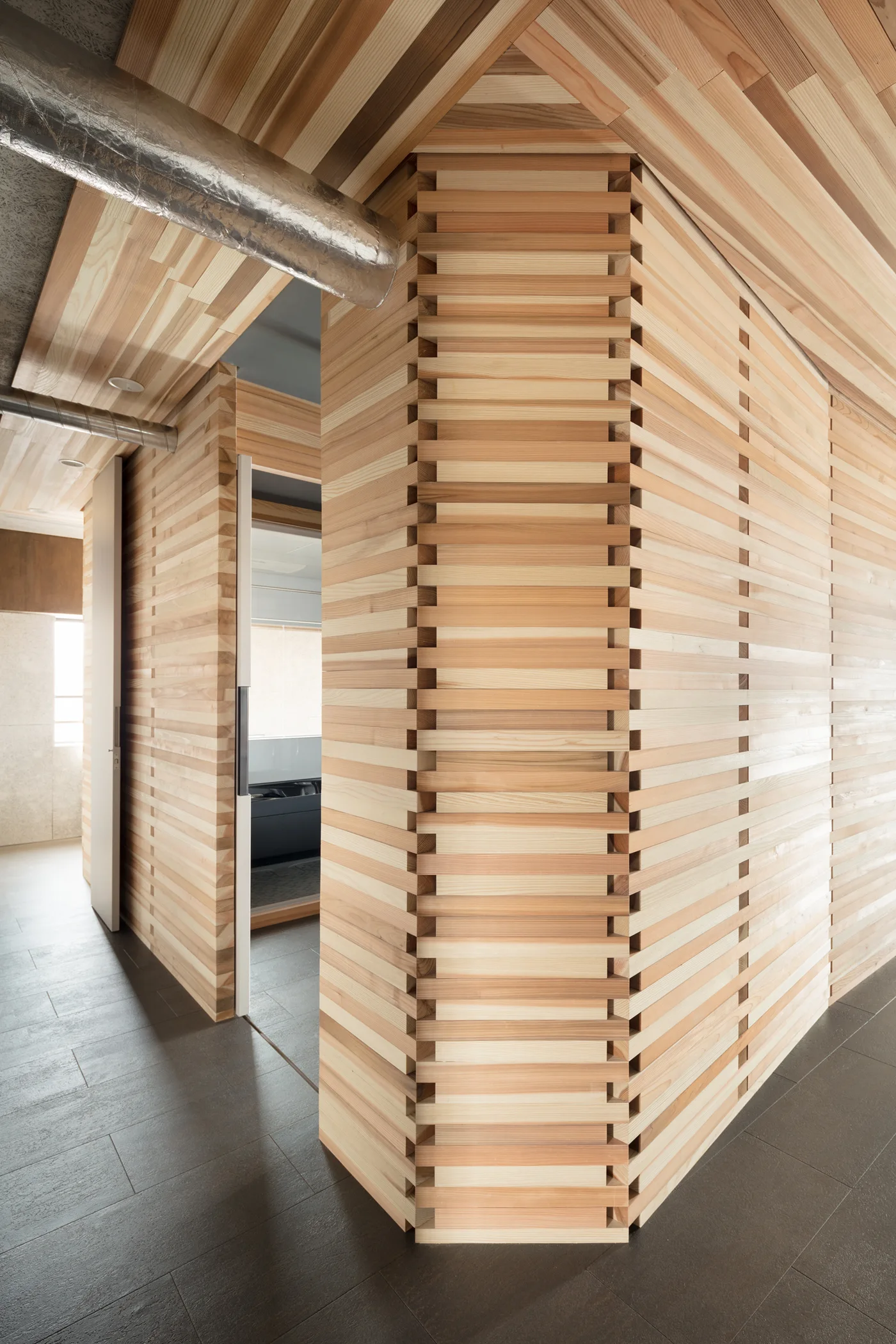 An architectural hallway featuring horizontal light wood slat wall panels, matching wood ceilings, and large-format dark gray floor tiles.