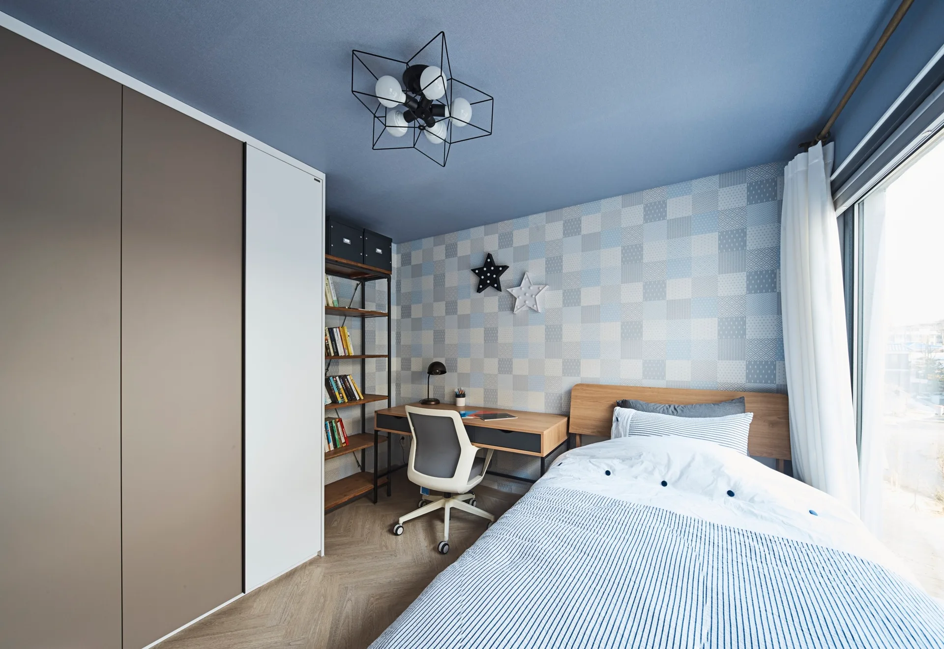 A modern bedroom features a bed with striped bedding, a wooden desk, a herringbone wood floor, and a painted blue ceiling.
