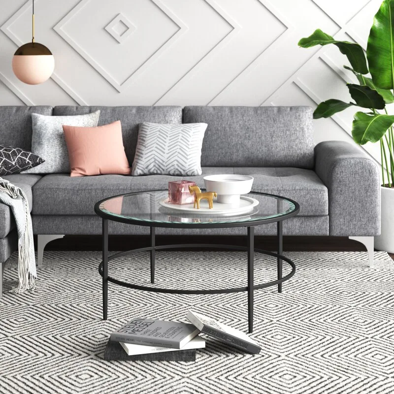 A grey sectional sofa and a round glass coffee table rest on a geometric rug against a white wall with geometric relief molding.