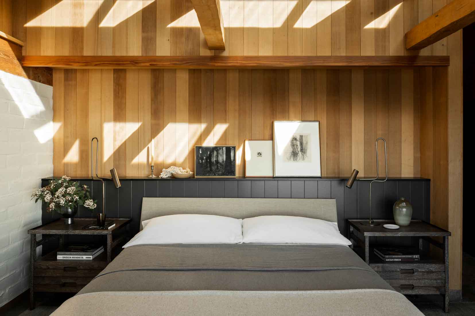 A bed with white and grey bedding sits against a black wood paneled half wall topped by a natural vertical wood plank wall.