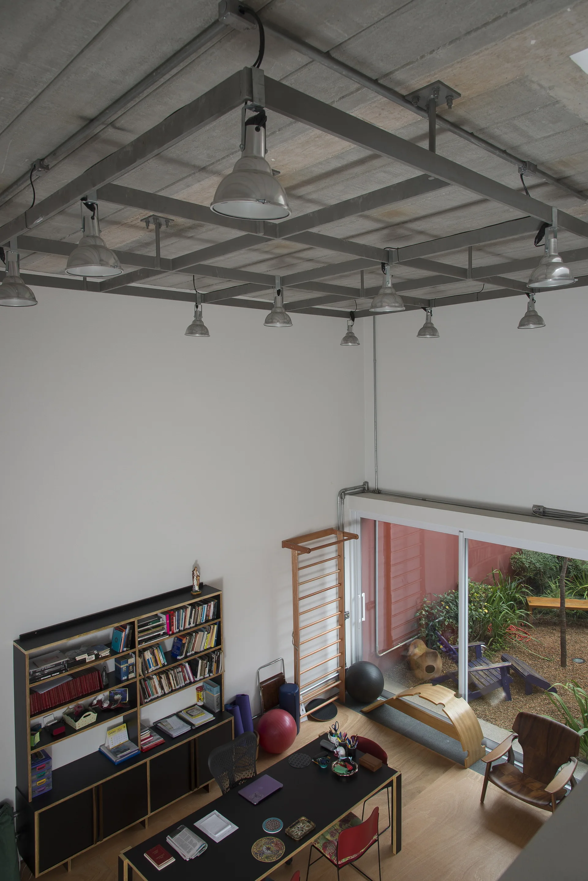 High-angle view of a home office with a black desk, wooden floors, tall white walls, workout wall bars, and a suspended metal ceiling grid with lights.