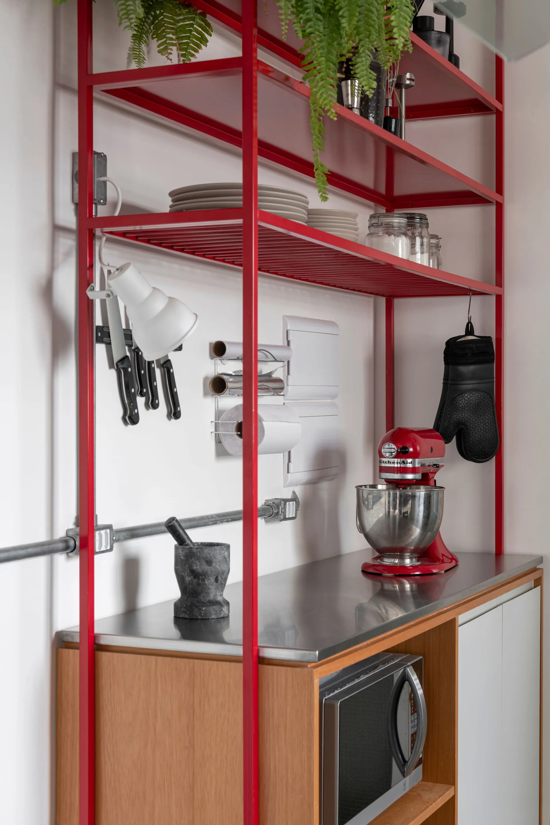 A red metal shelving unit stands over a stainless steel countertop with a matching red stand mixer and exposed wall conduit.