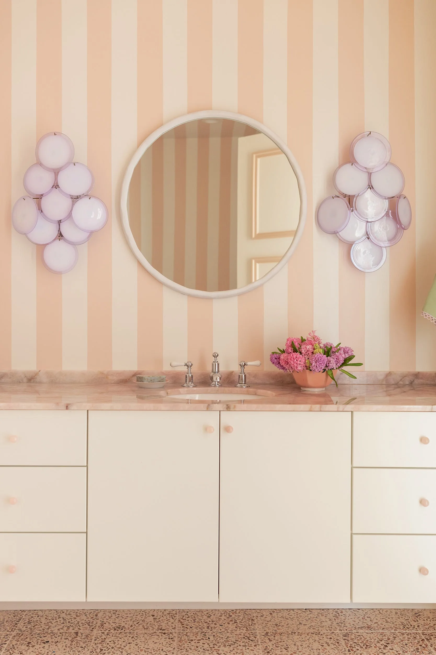 A bathroom vanity features a pink marble countertop, a round white mirror, lavender glass sconces, and peach and cream striped wallpaper.