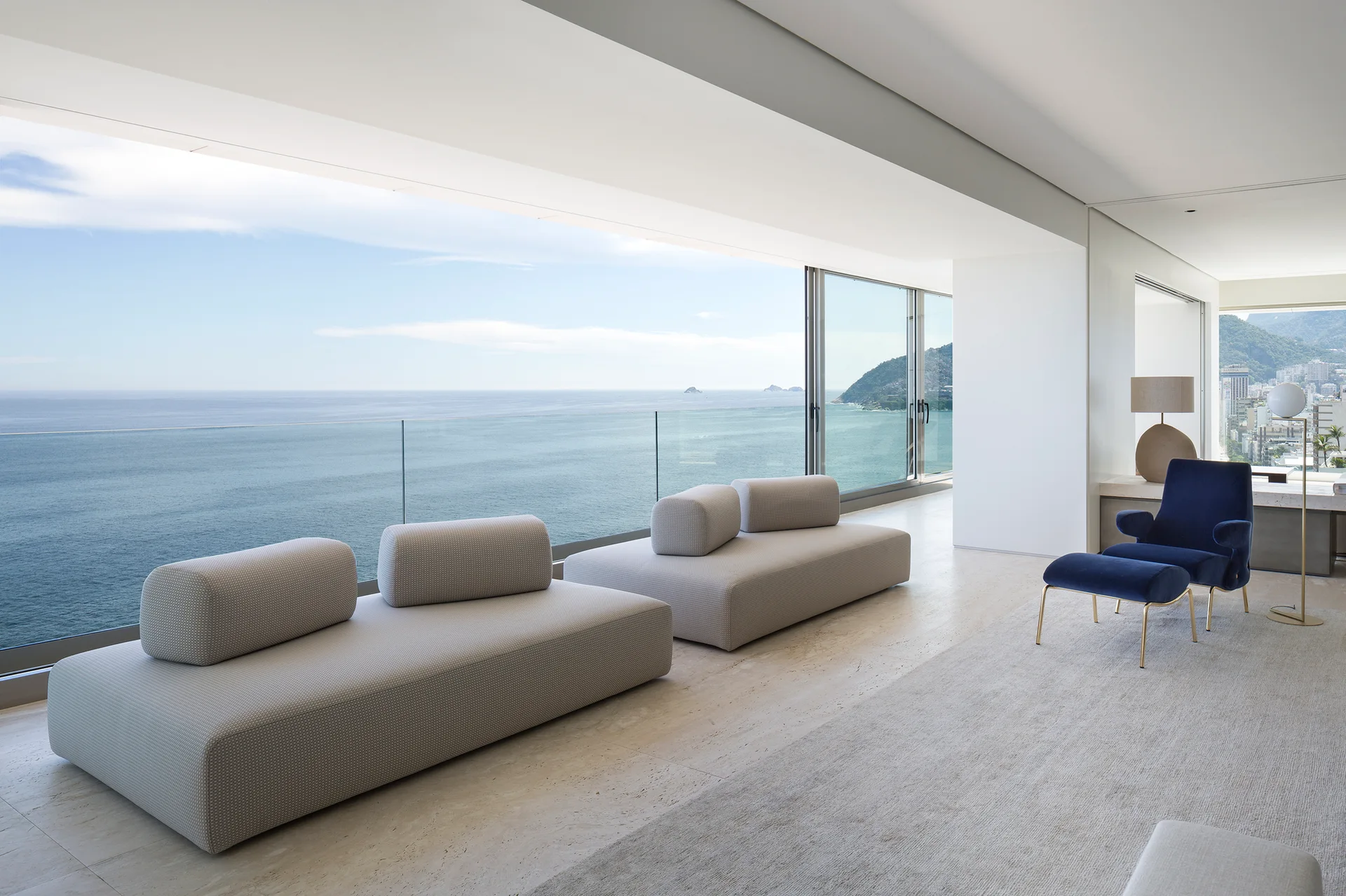 Two low-profile modular sofas and a navy blue velvet armchair face a large glass window wall with an expansive ocean view.