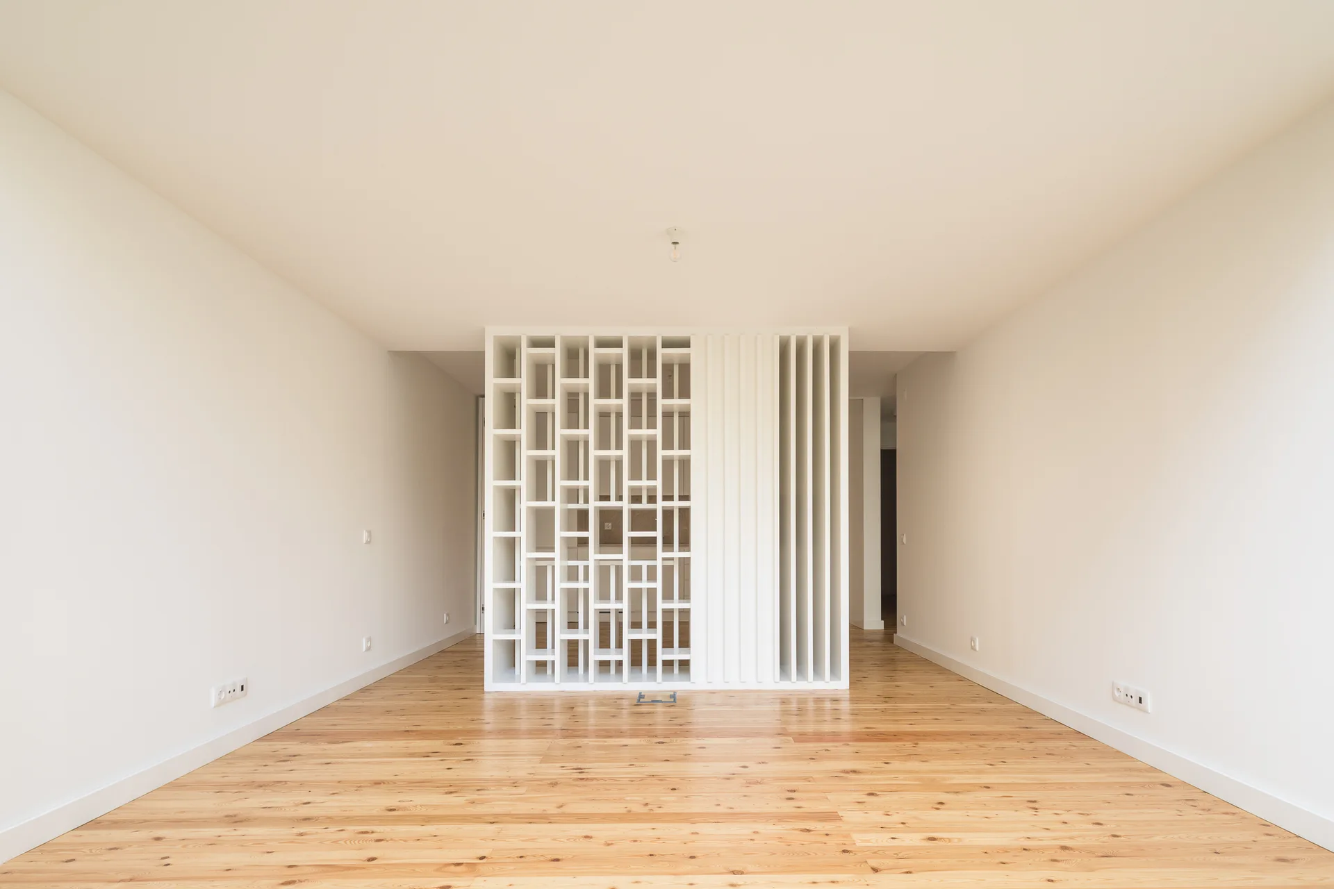 An empty minimalist room features natural light wood plank flooring and a central white geometric grid screen divider.