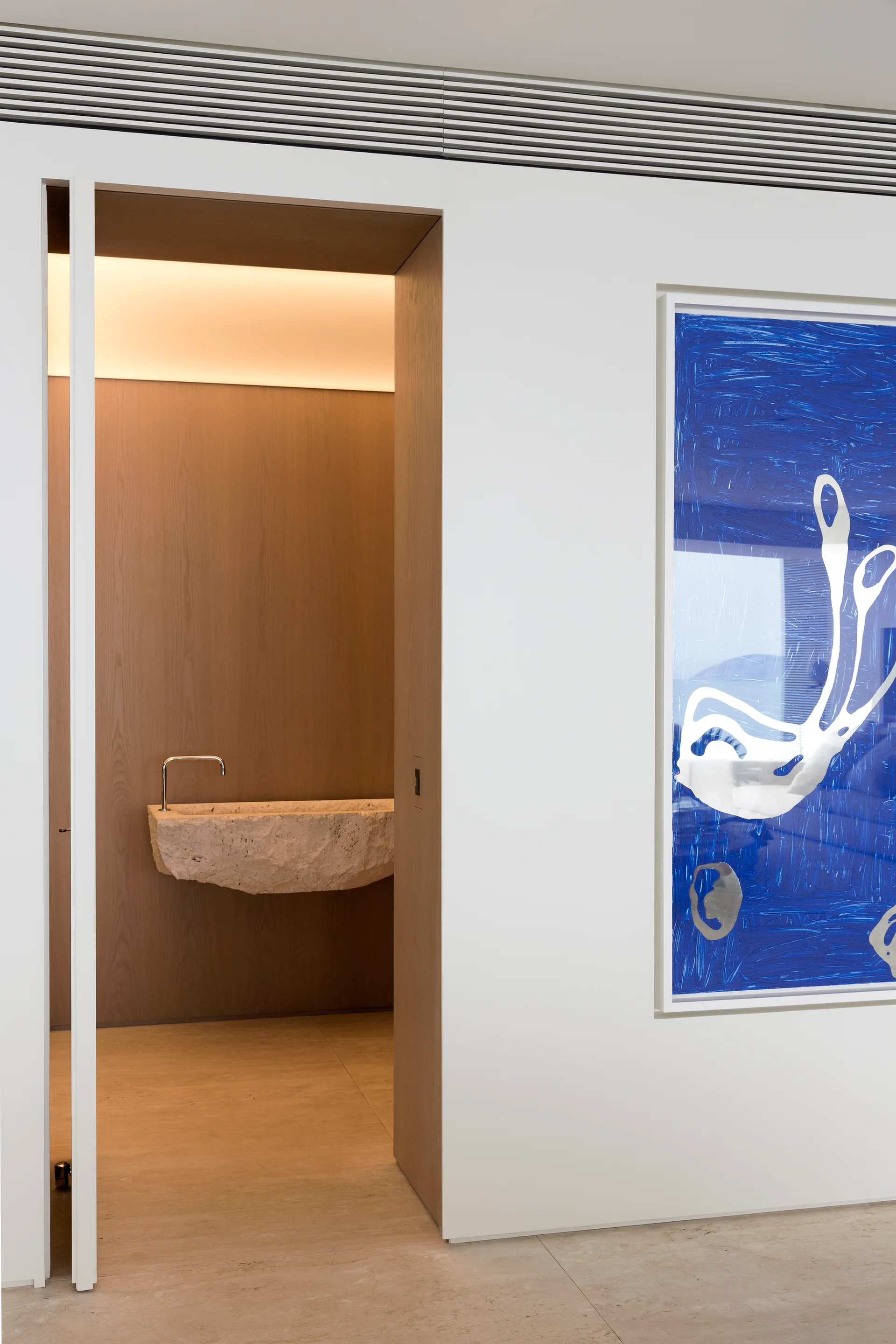 A minimalist doorway opens to a wood-paneled powder room with a rough-hewn stone basin, next to a white wall featuring blue abstract artwork.