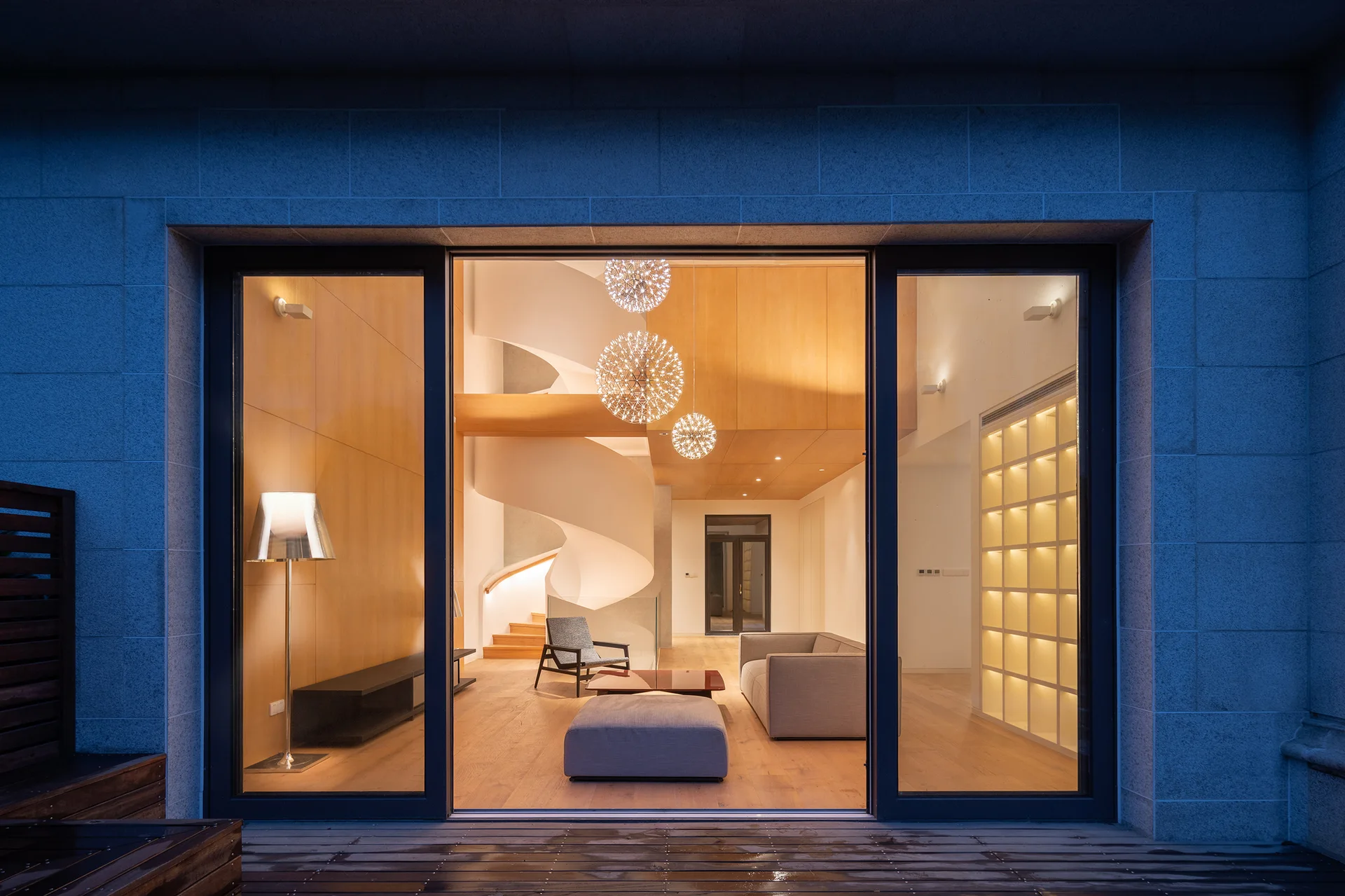A modern living room with a white spiral staircase, grey sofa, and illuminated shelving seen through large sliding glass doors.