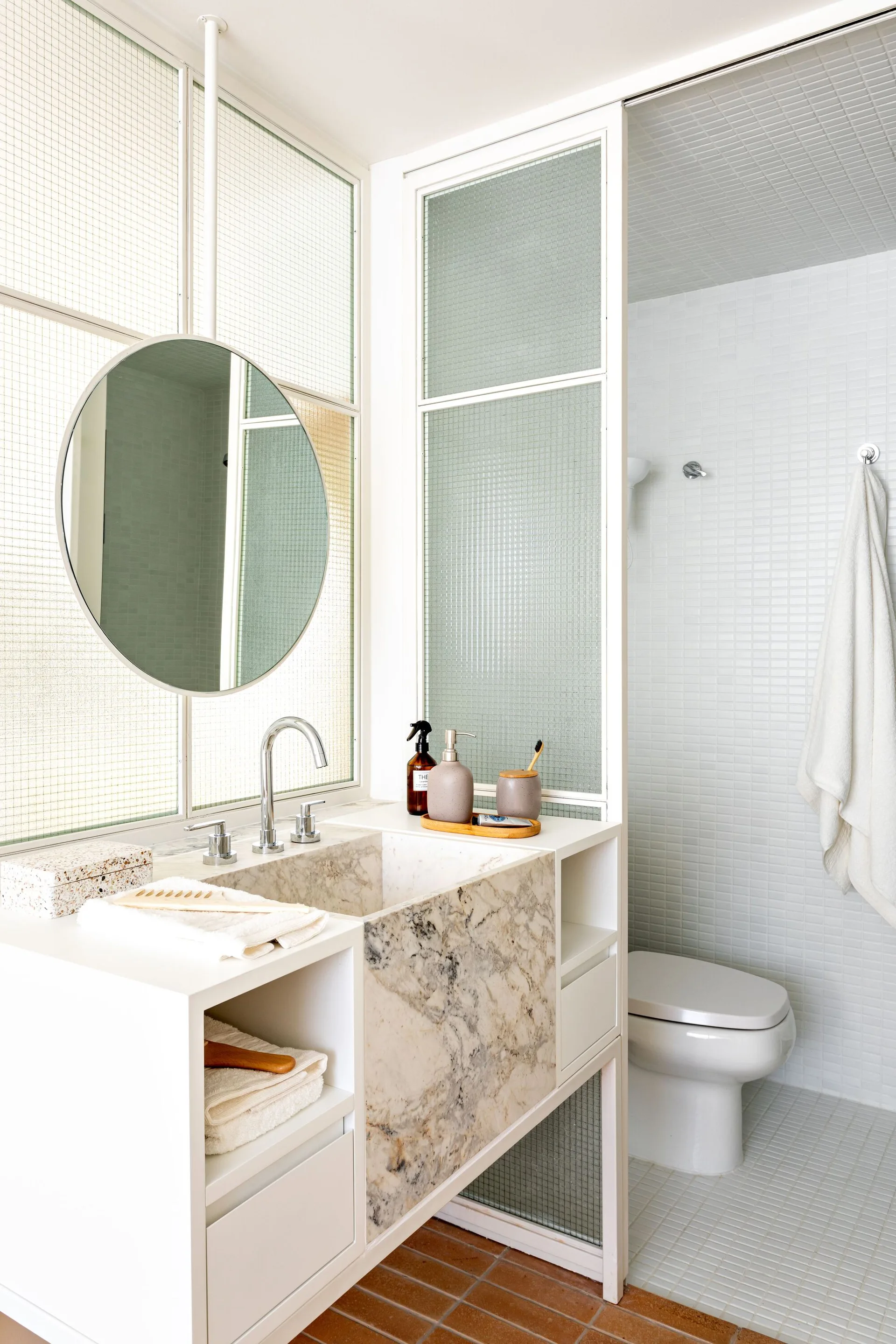 A white bathroom features a marble-front vanity, a round wall mirror, and a grid-framed glass partition dividing the sink from the toilet.