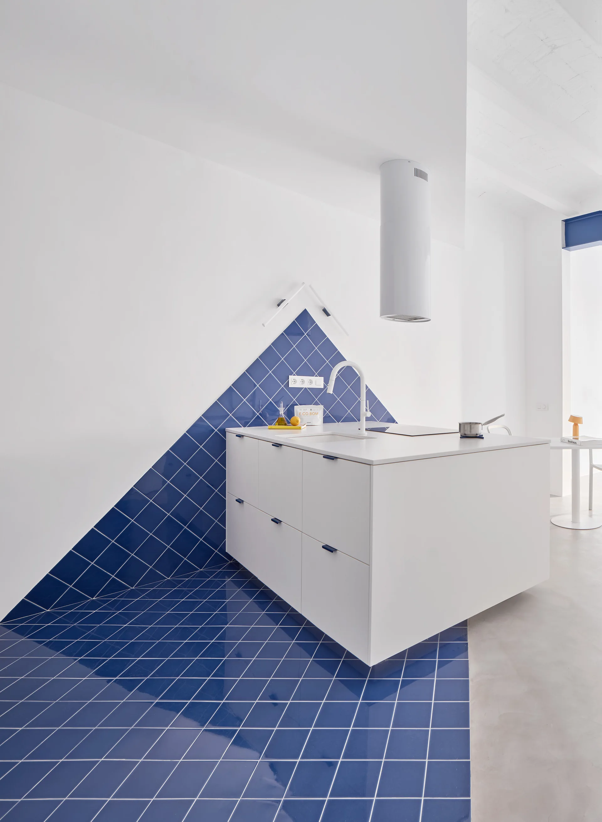 A white minimalist kitchen island sits on a bright blue tiled floor that climbs the back wall in a matching triangular backsplash pattern.