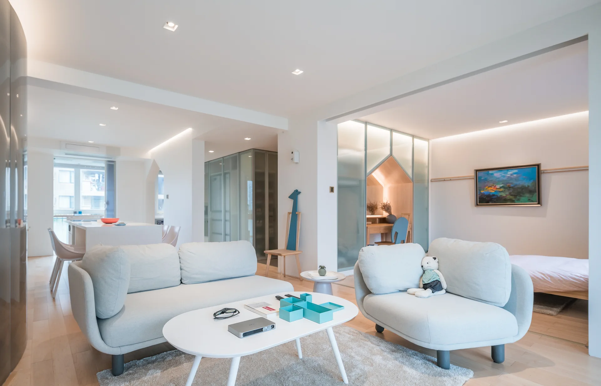 A bright, open-plan living room features pale rounded sofas, an oval white coffee table, light hardwood floors, and a translucent glass room partition.