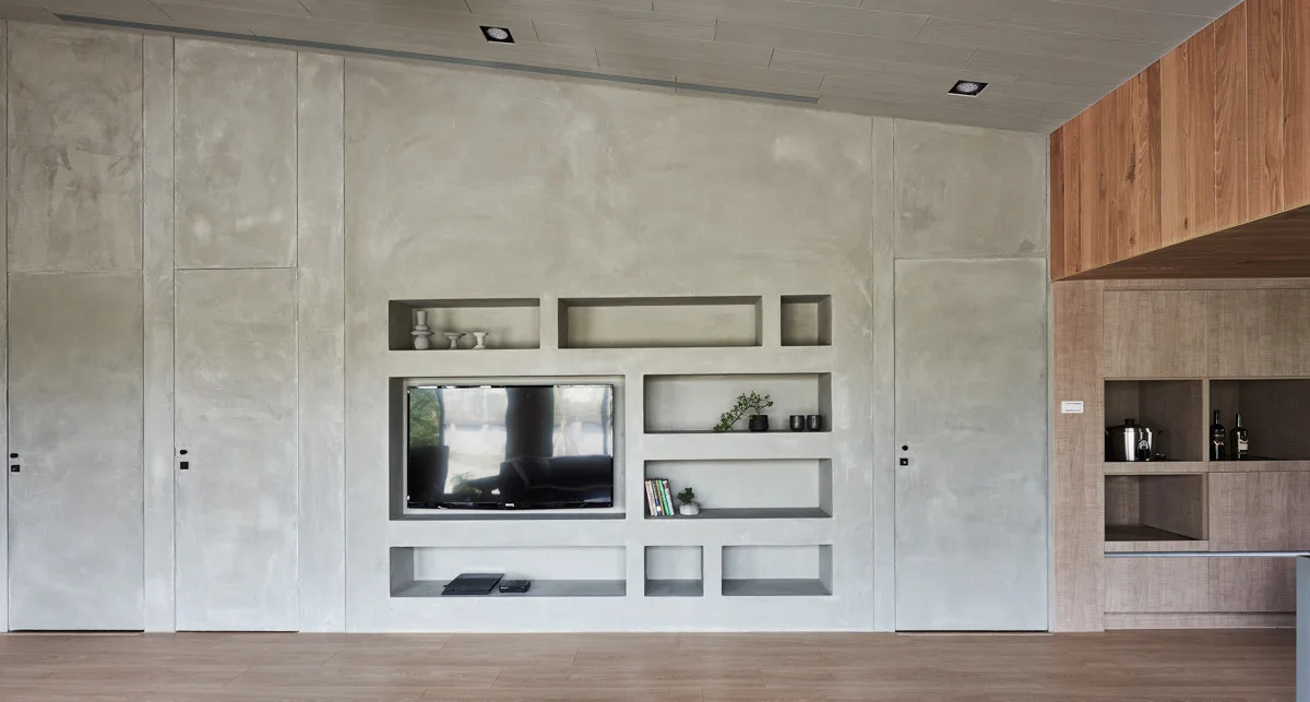 A minimalist grey concrete-look wall features recessed geometric shelving, an integrated television, and two flush hidden doors.