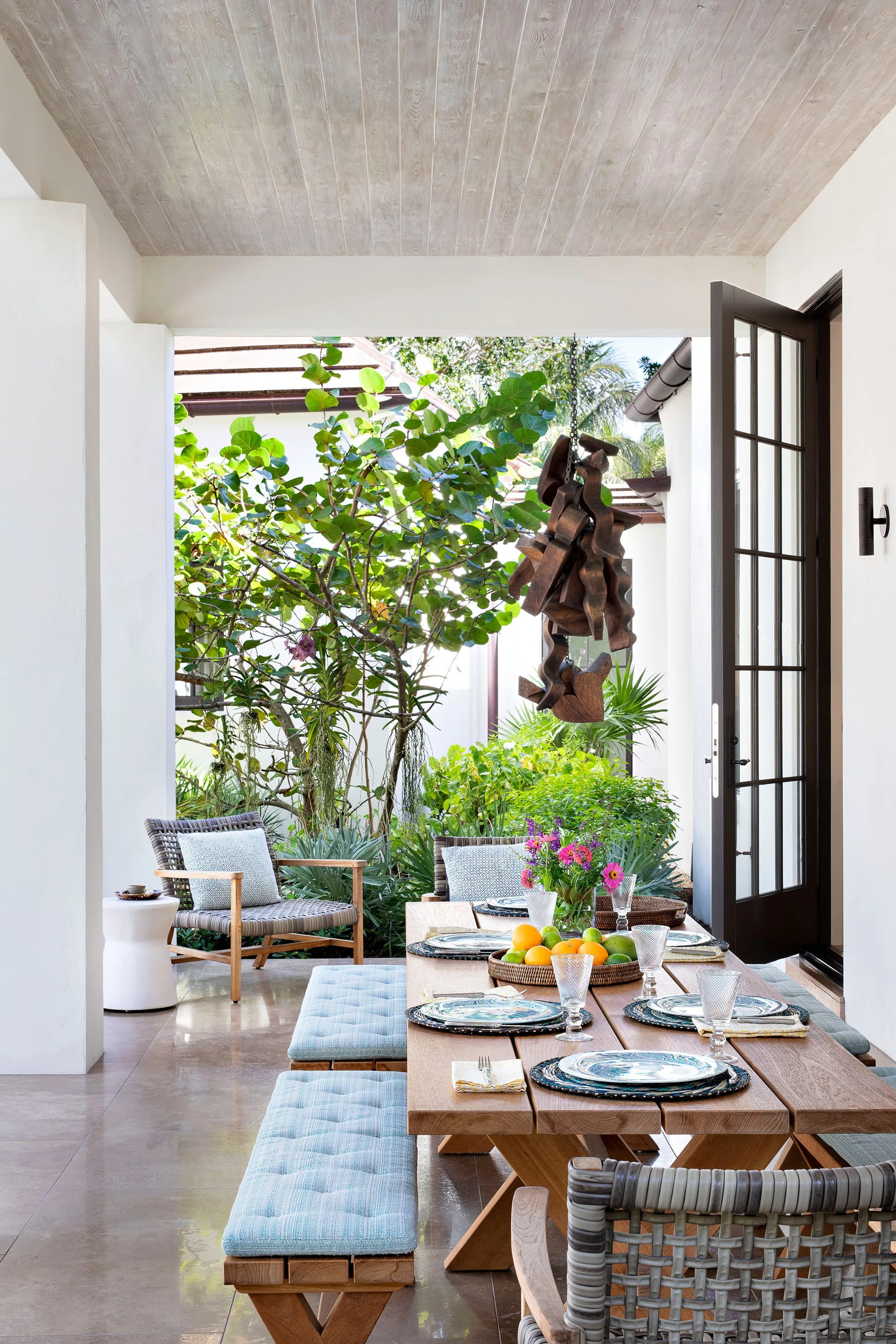 A covered patio features a wooden dining table with light blue cushioned benches, woven chairs, a wood-plank ceiling, and a view of a lush green garden.
