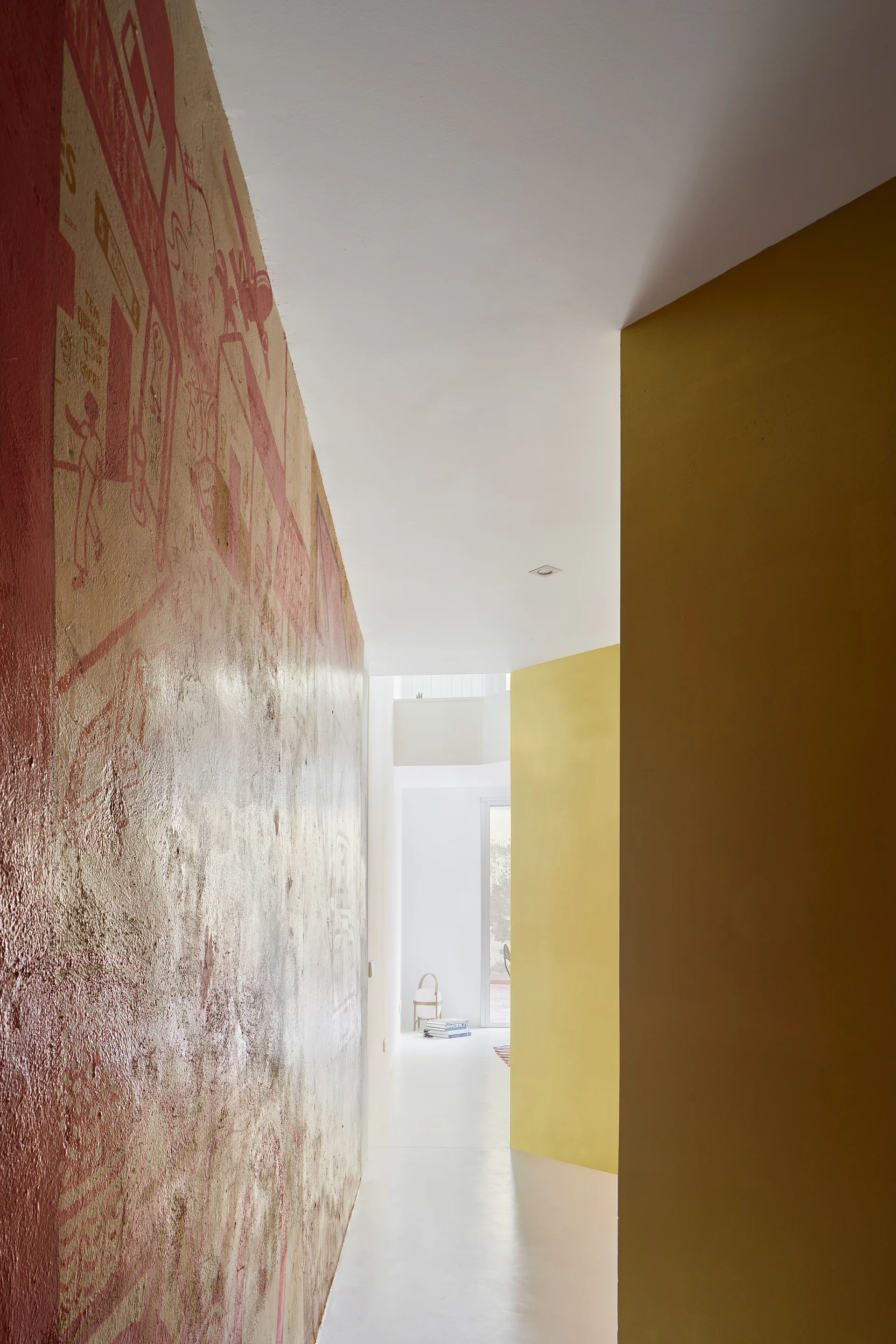 A minimalist hallway features a textured red graphic mural wall opposite a smooth yellow accent wall, leading to a bright white space in the distance.