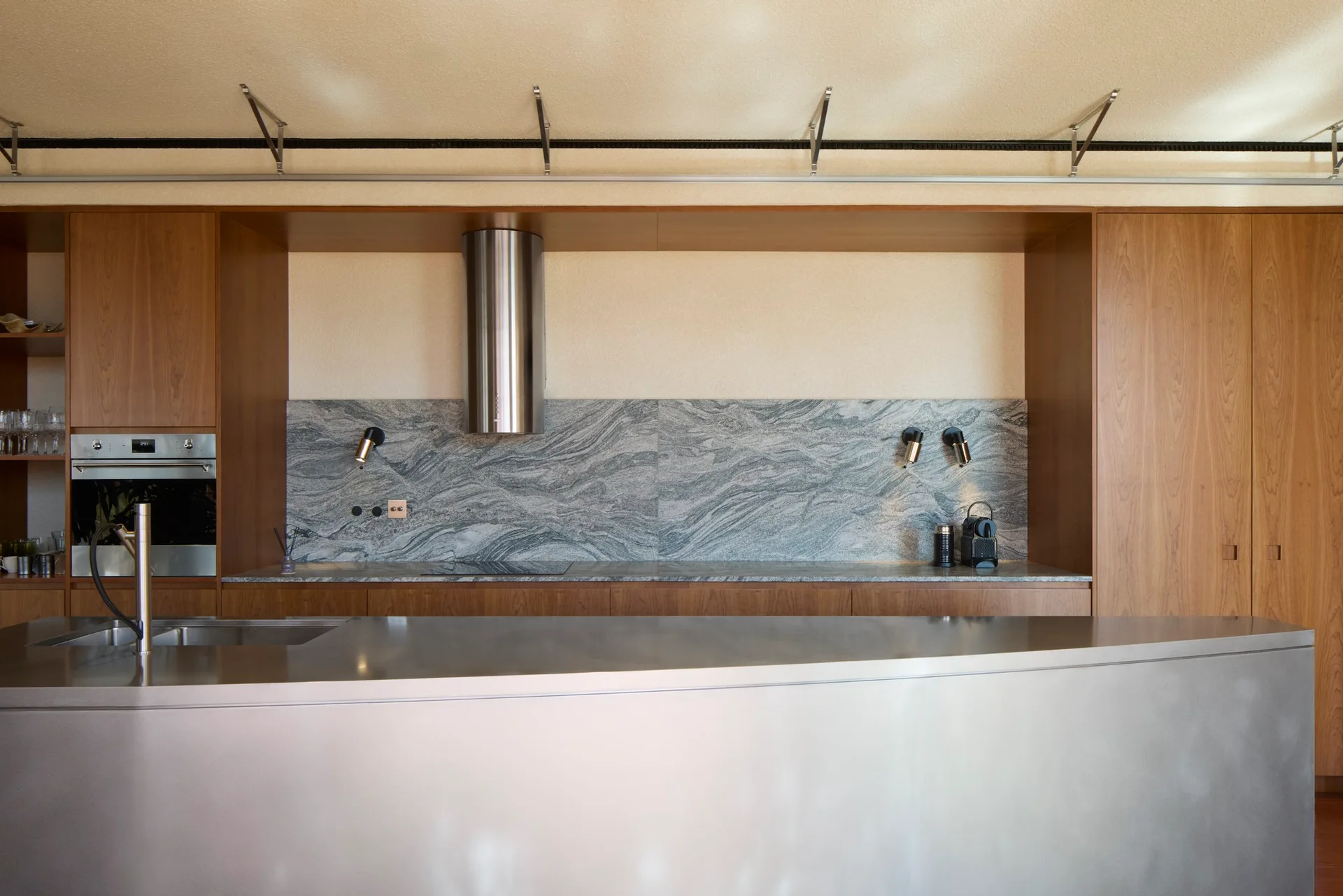 A minimalist kitchen features warm wood cabinetry, a grey veined stone backsplash, a cylinder range hood, and a reflective stainless steel island.