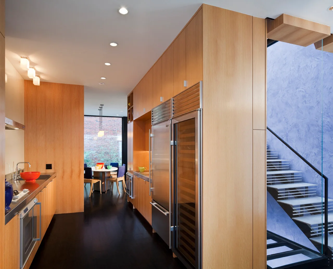 A modern galley kitchen with light wood cabinets, stainless steel refrigerator, and an open staircase featuring a blue textured wall.