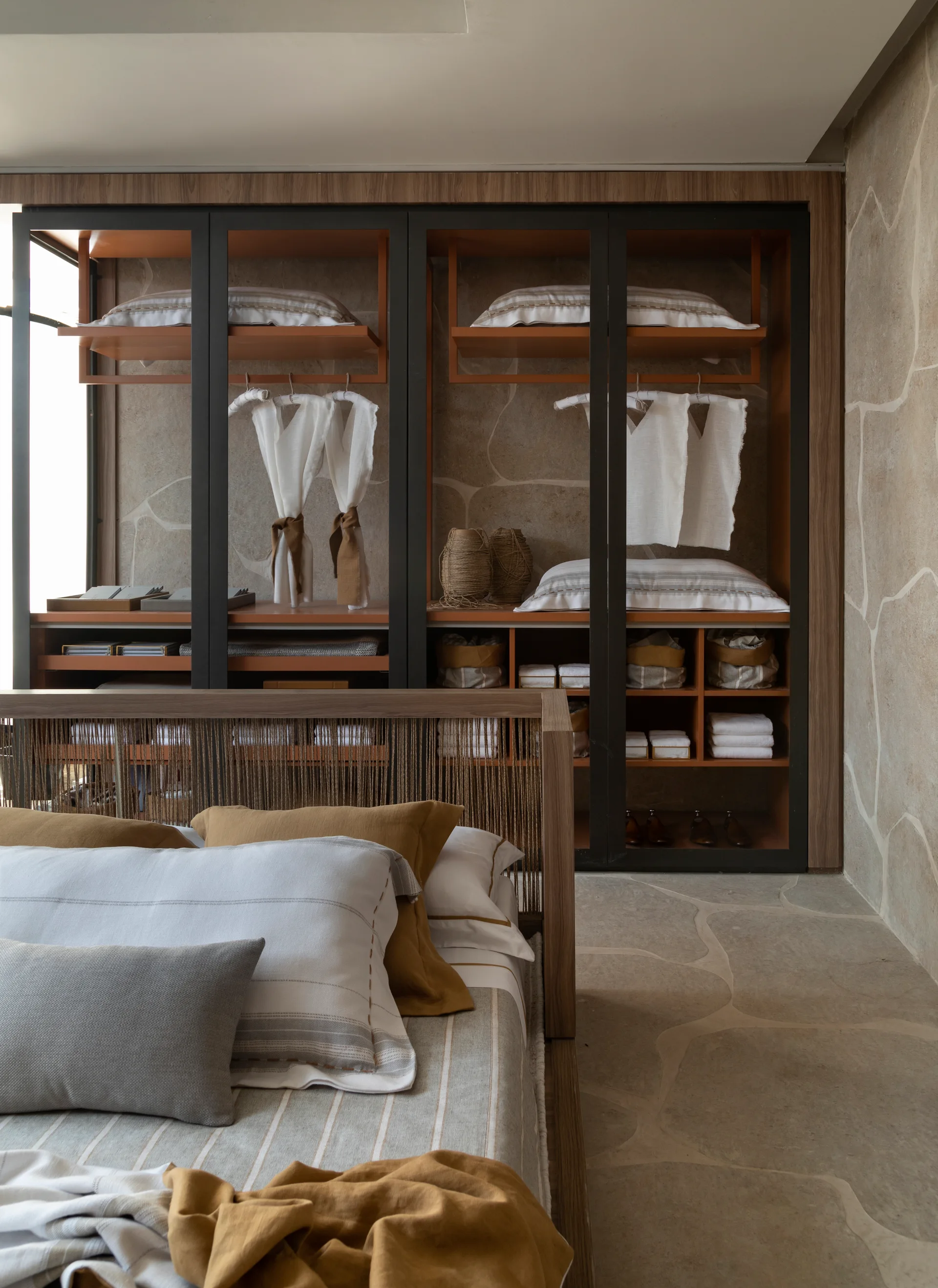 A modern bedroom features a wooden bed with striped linens next to a built-in dark wood closet with glass doors and a stone-clad wall.