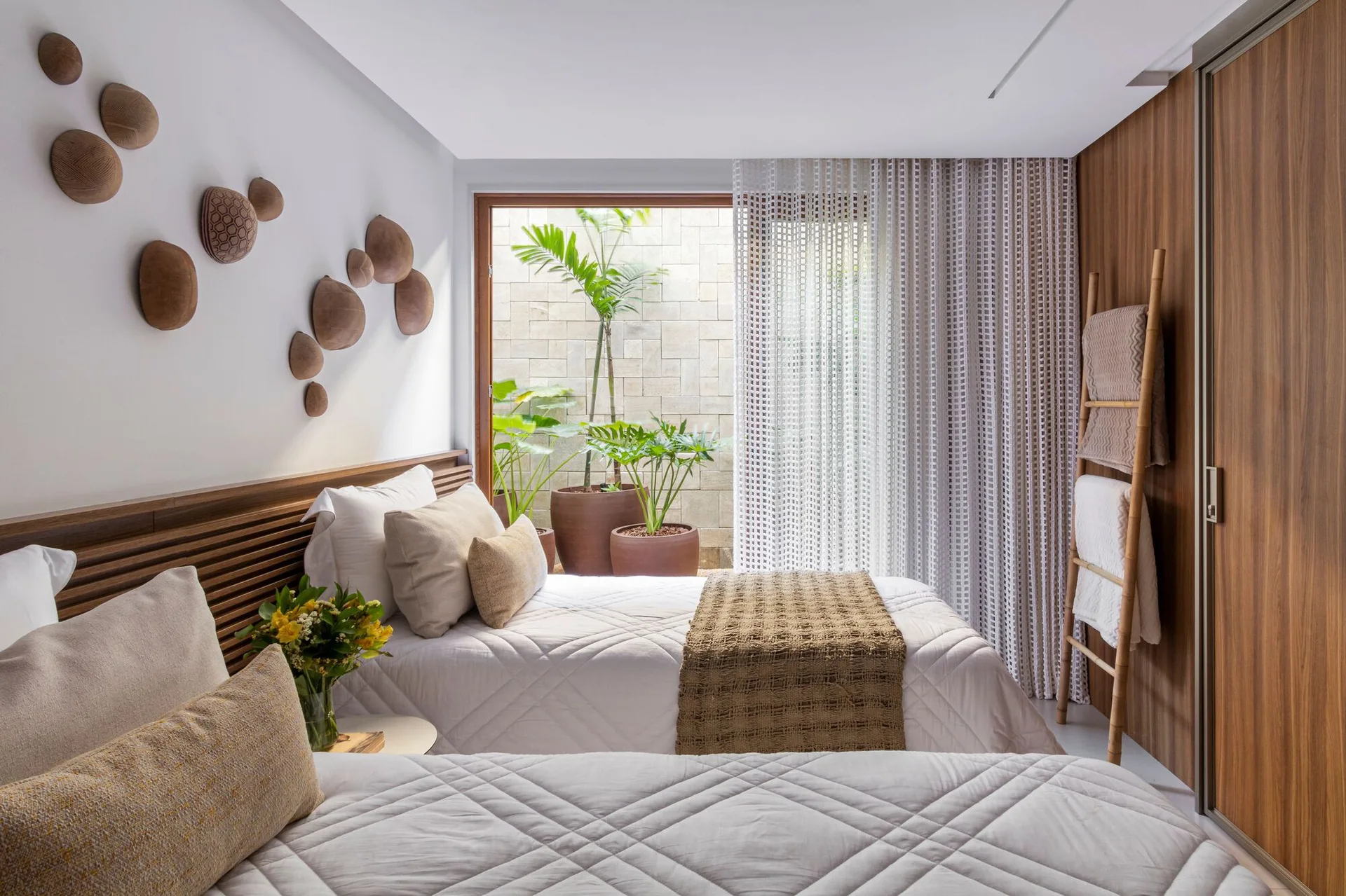 Two twin beds with white quilted blankets rest against a slatted wood headboard, opening to a private patio with potted plants.