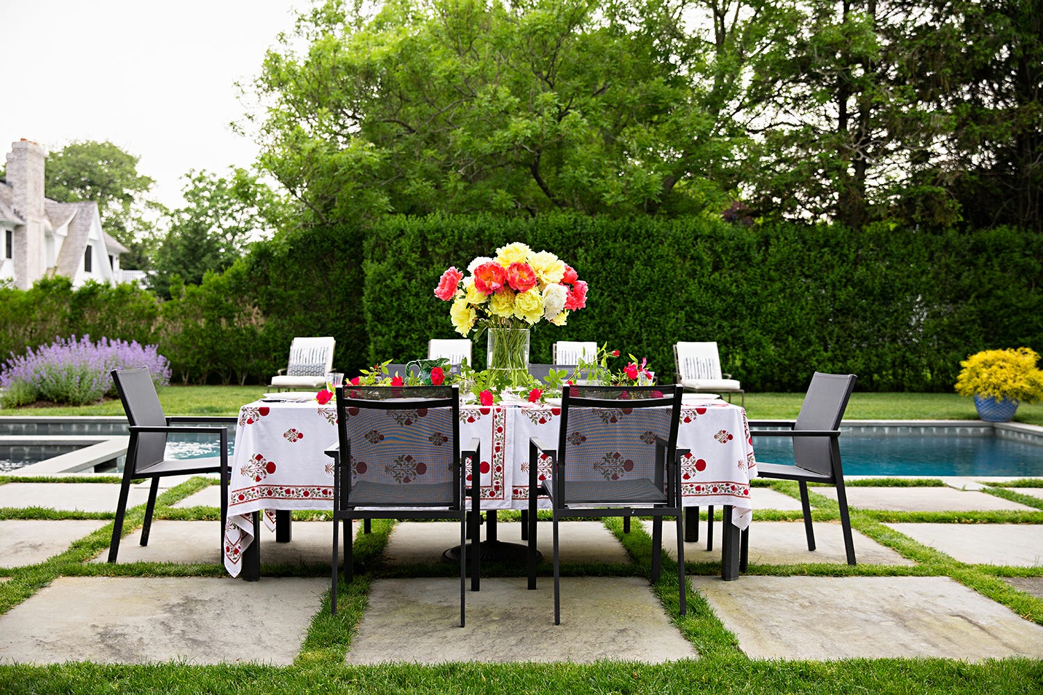 An outdoor dining table with a red patterned tablecloth and floral centerpiece sits on stone pavers next to a swimming pool and tall hedge.