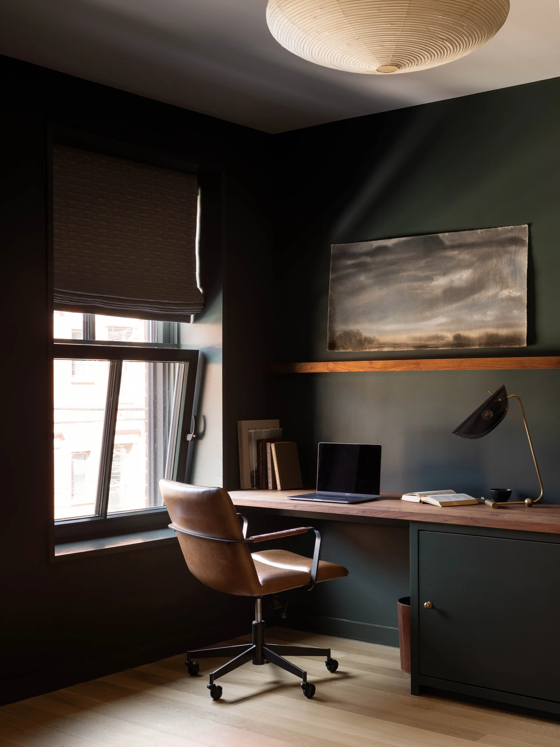 A dark green home office features a natural wood desk, a brown leather swivel chair, a window with a Roman shade, and a brass desk lamp.