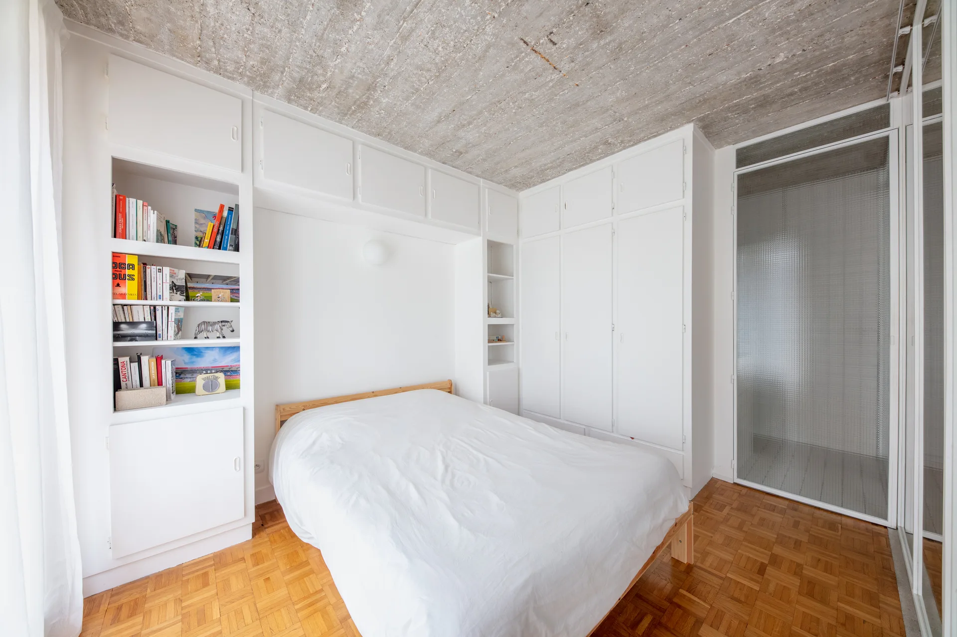 A minimalist bedroom features a wooden bed with white bedding, surrounded by floor-to-ceiling white storage units under a textured concrete ceiling.