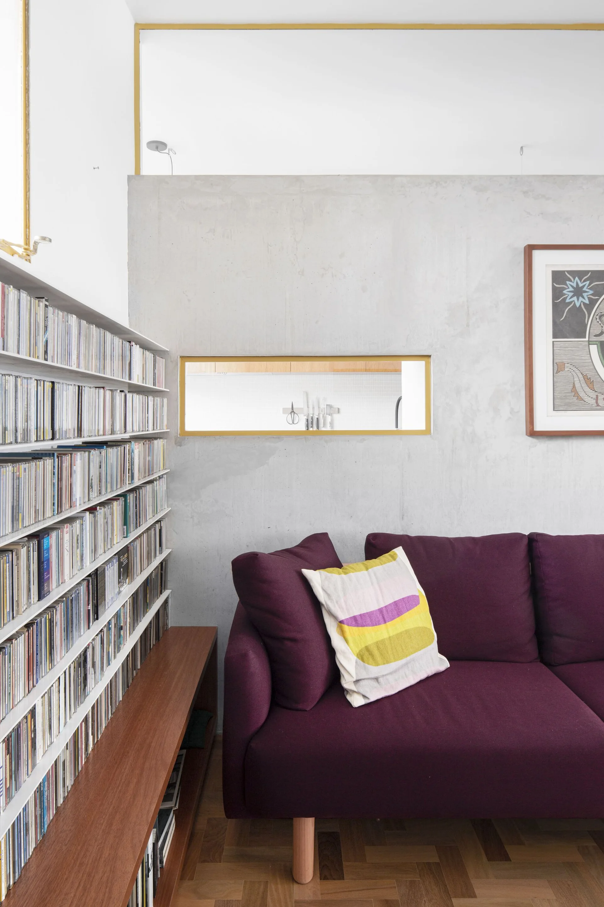 A purple sofa sits against a smooth grey concrete wall featuring a small rectangular pass-through window and a tall media shelf filled with cases.