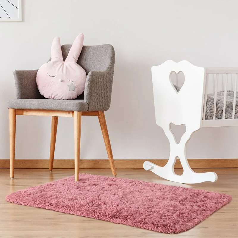 A nursery features a white wooden cradle with heart cutouts, a grey upholstered armchair with a pink bunny pillow, and a fluffy pink rug on wood floors.
