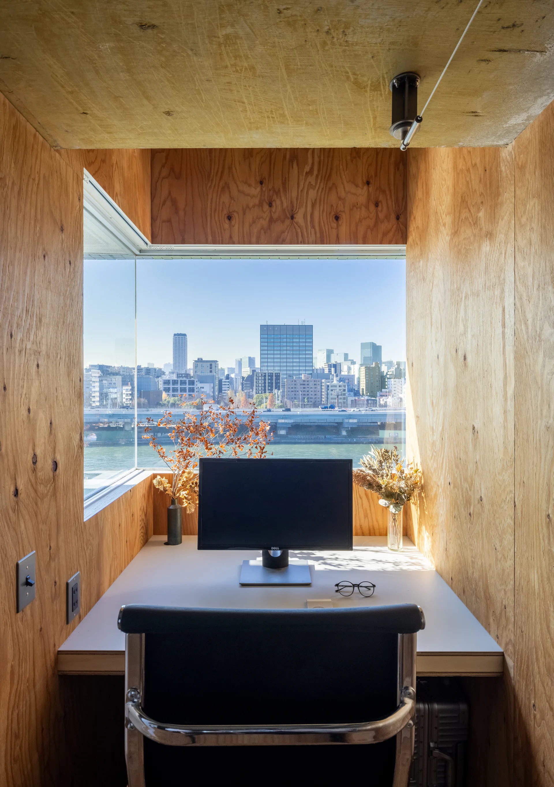 A compact built-in desk constructed from plywood features a computer monitor and a large window looking out onto a river and city skyline.