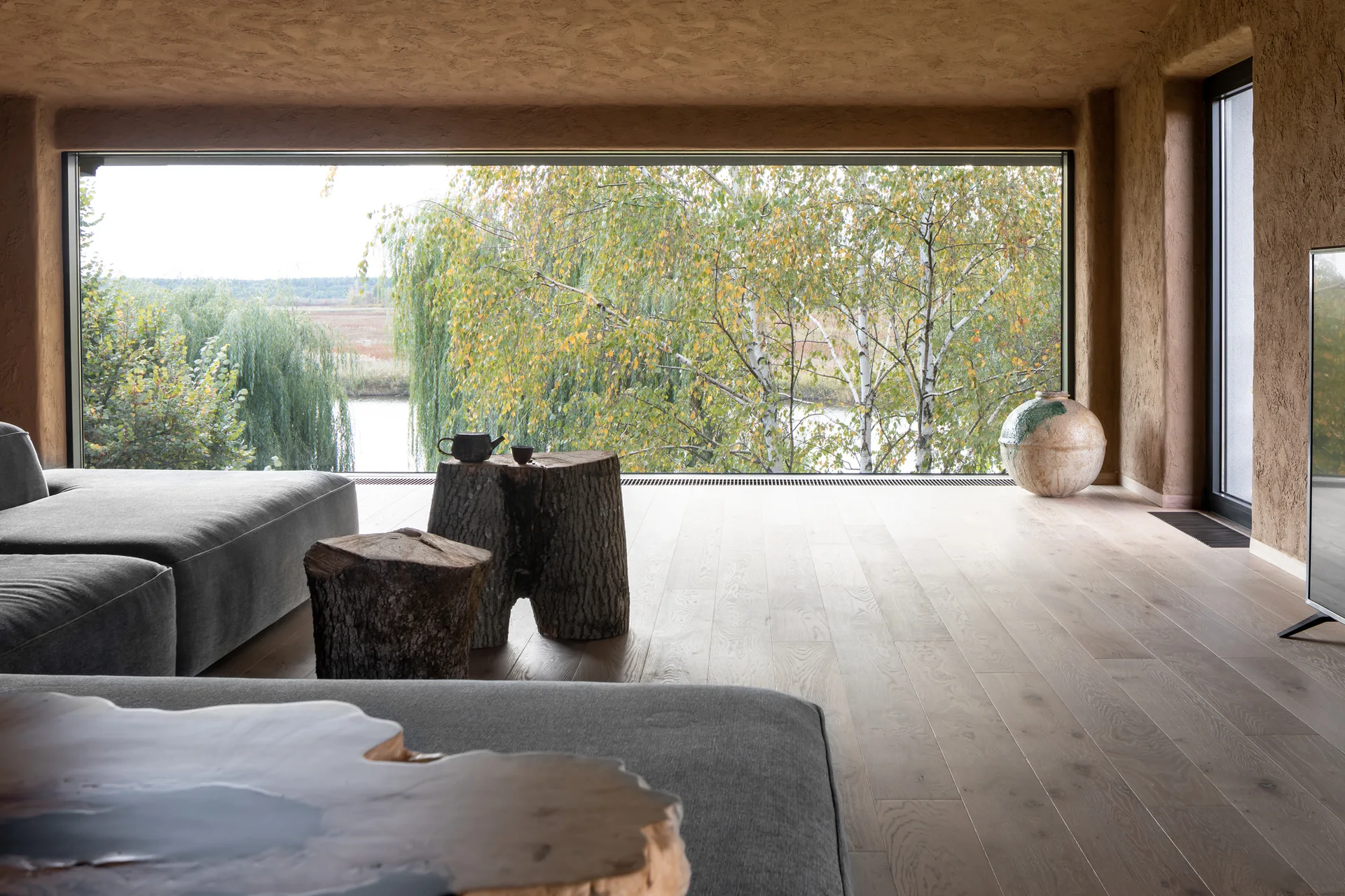 A minimalist living room features a grey sectional sofa, rustic log side tables, and a large panoramic window displaying trees outside.