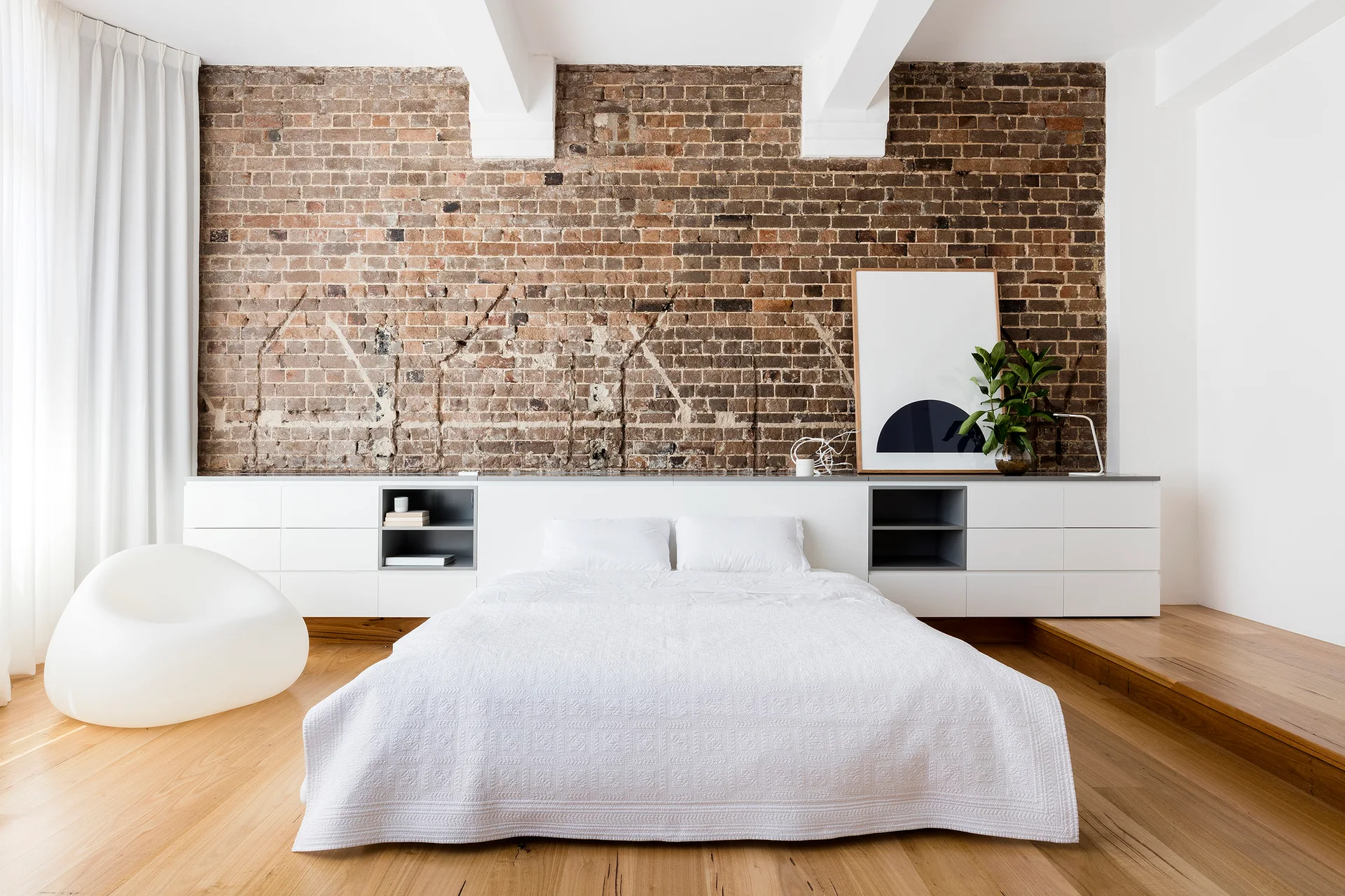 A low white bed sits in front of a long white built-in storage cabinet and a rustic exposed brick wall in a bright modern bedroom.