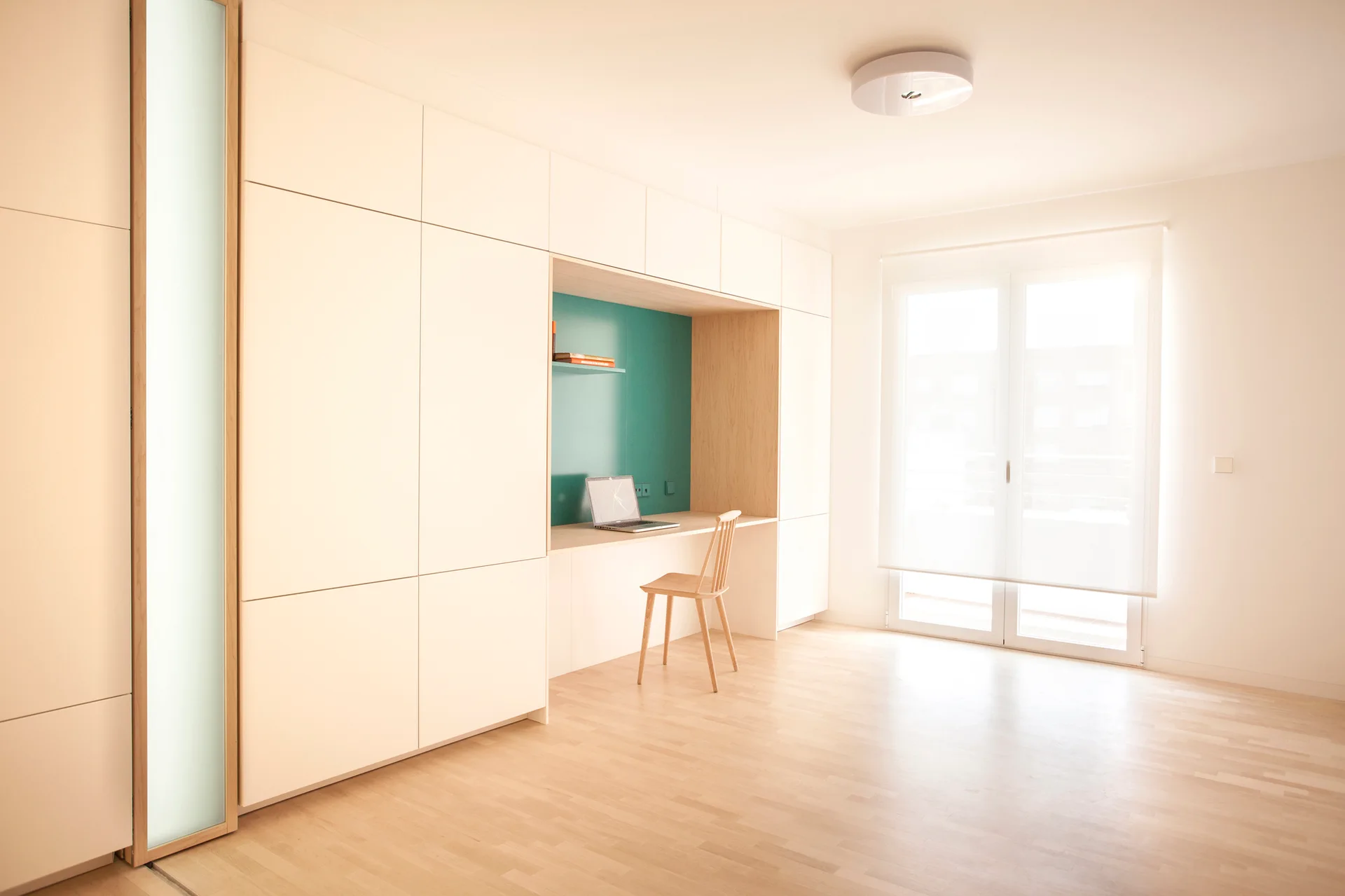 A minimalist home office featuring a white built-in storage cabinet wall with an integrated wooden desk nook and a teal accent panel.