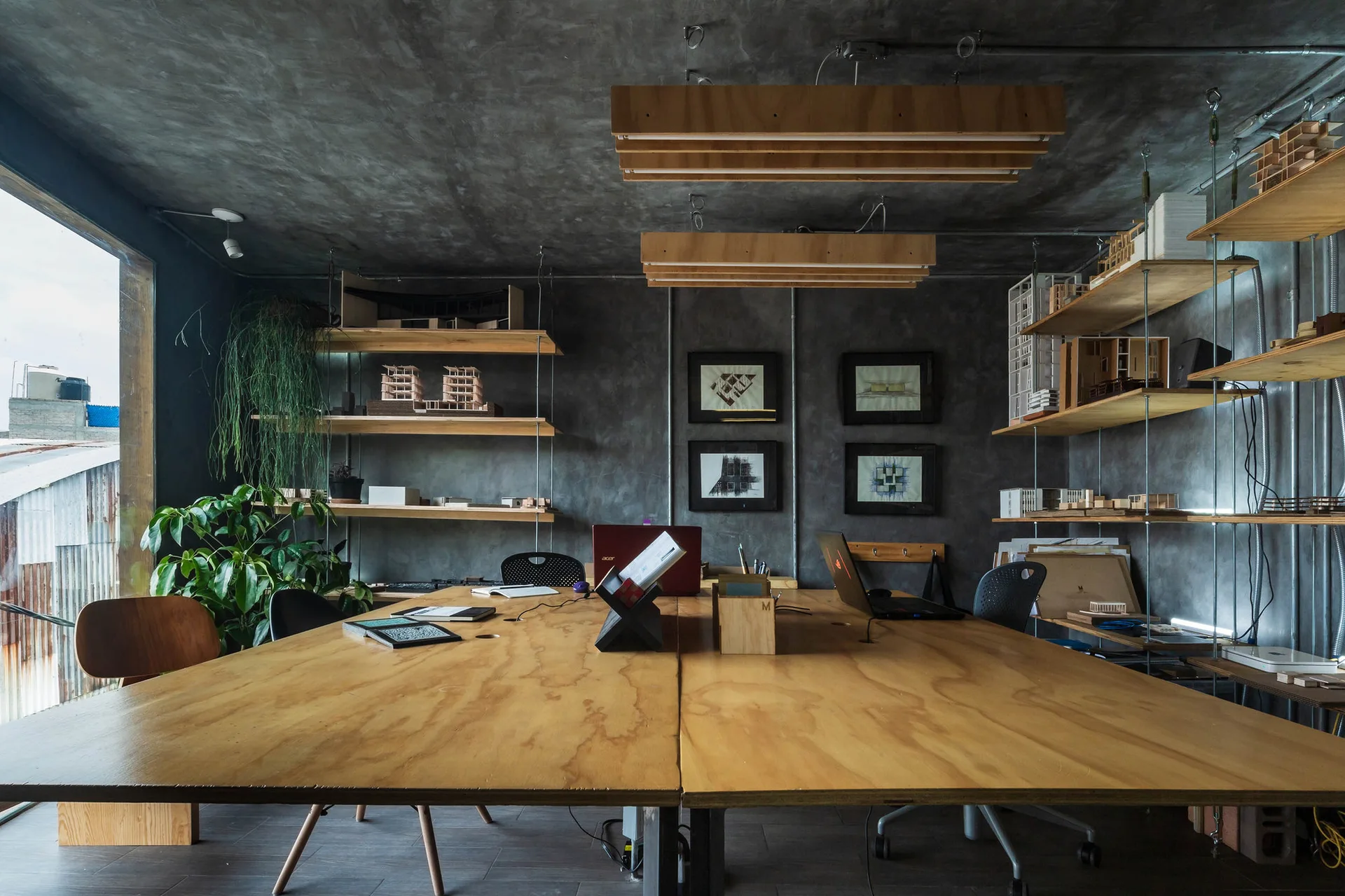 An industrial studio office features a large plywood desk, raw concrete walls, suspended wood shelves with miniature models, and custom linear lights.