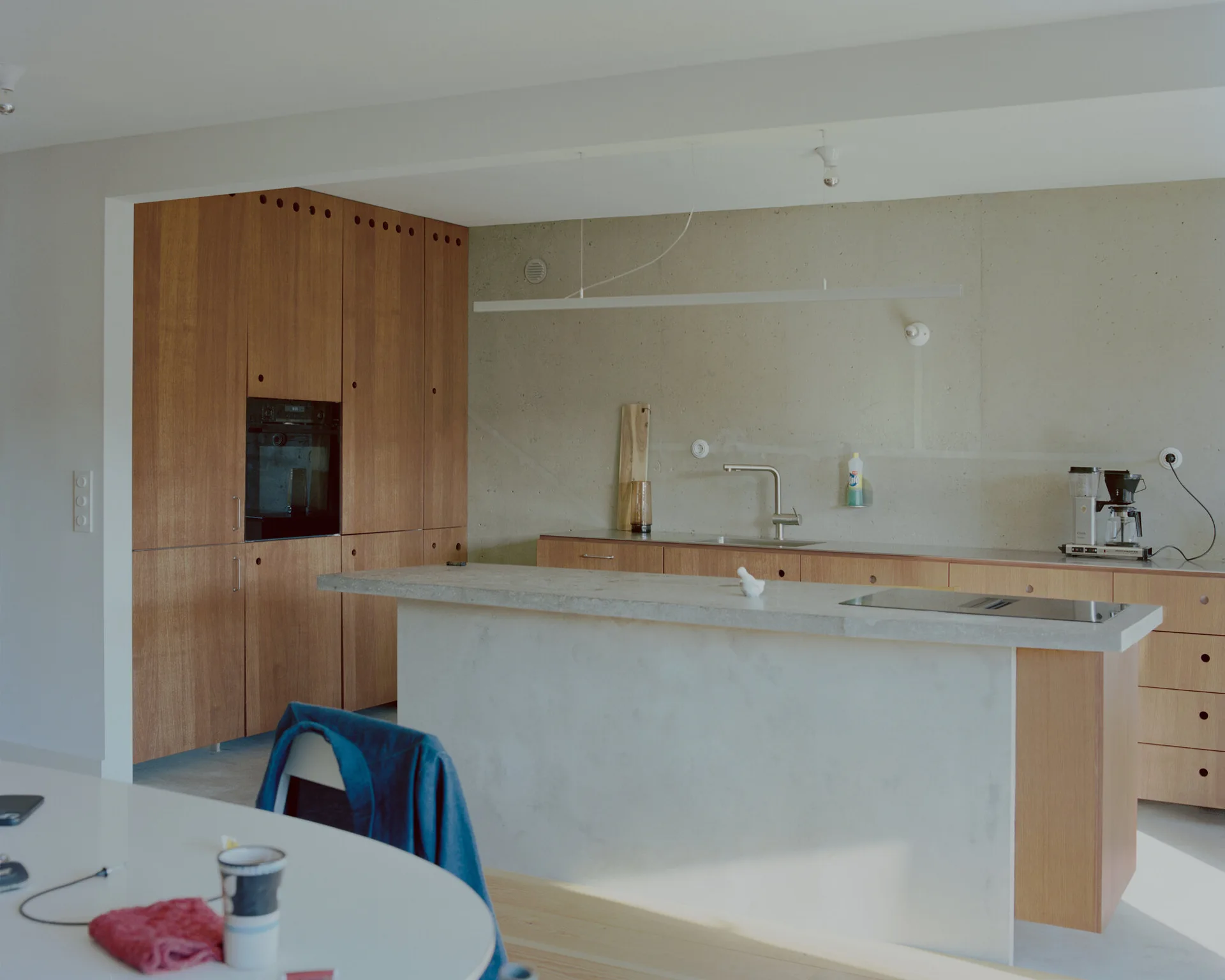 A minimalist kitchen features tall warm wood cabinets, a raw concrete wall, and a grey concrete island with a stone countertop.