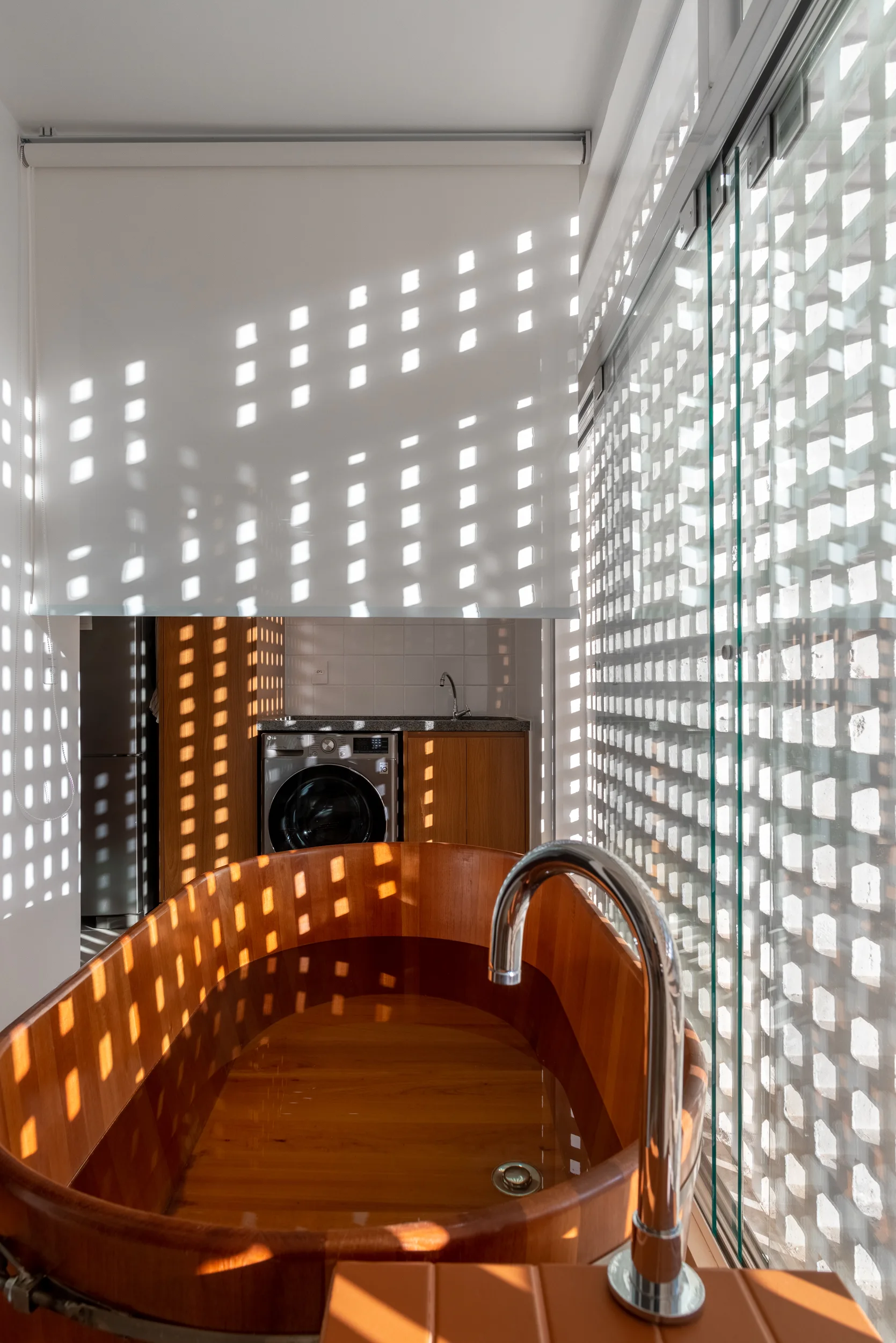 A wooden soaking tub sits in a bathroom where natural light filters through a lattice screen wall, casting checkered shadows on a white roller shade.