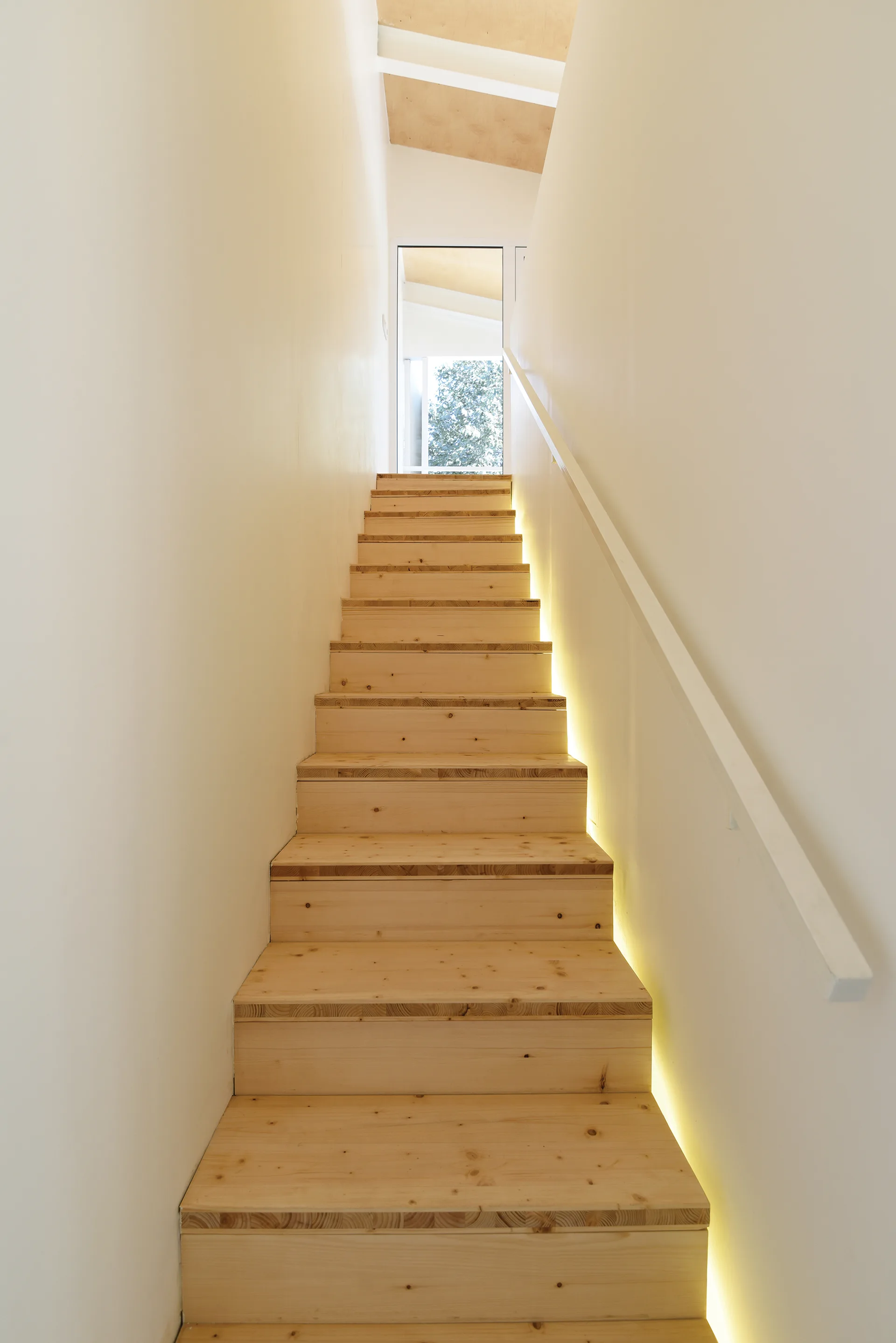 A straight wooden staircase with light pine steps is flanked by white walls and illuminated by a warm LED light strip along the right edge.
