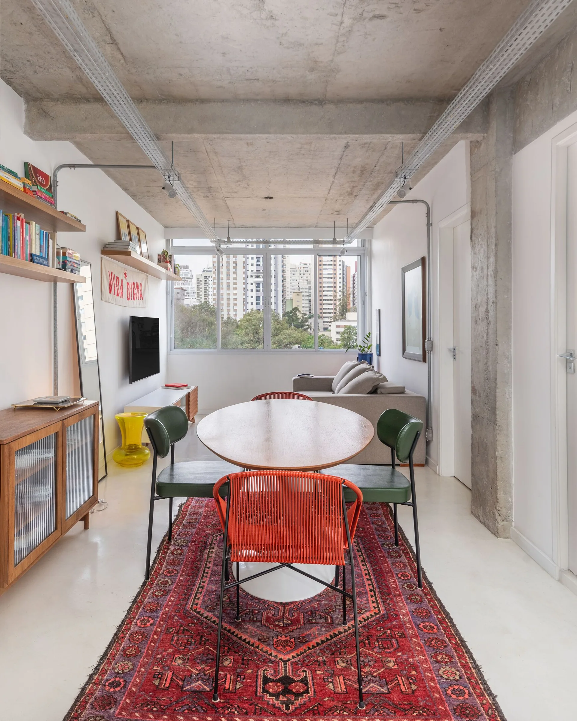 A wooden dining table with green and orange chairs sits on an ornate red rug beneath an exposed concrete ceiling in a bright apartment.