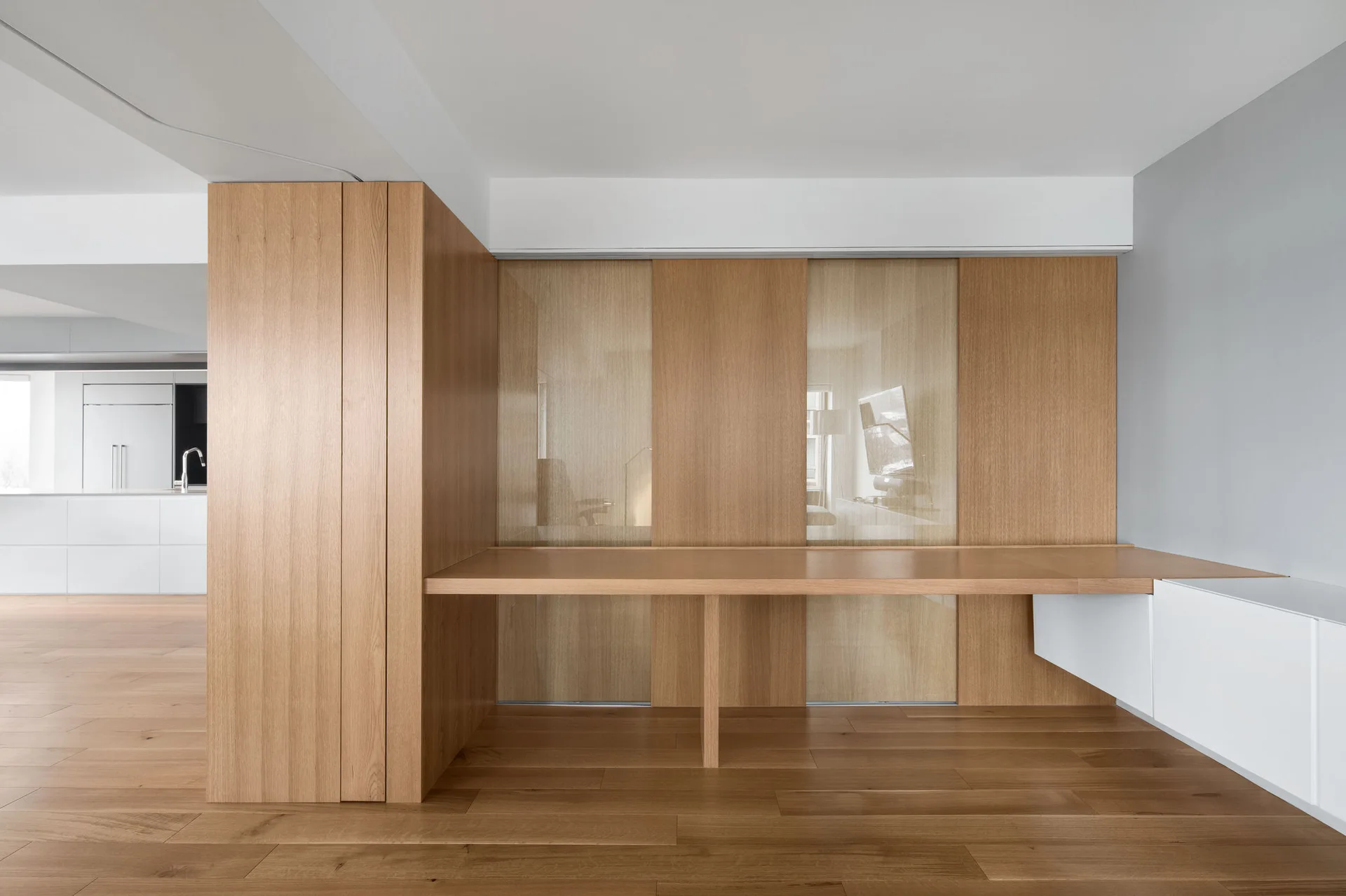 A minimalist room featuring a long light wood built-in desk integrated with matching wall paneling and a low white cabinet.