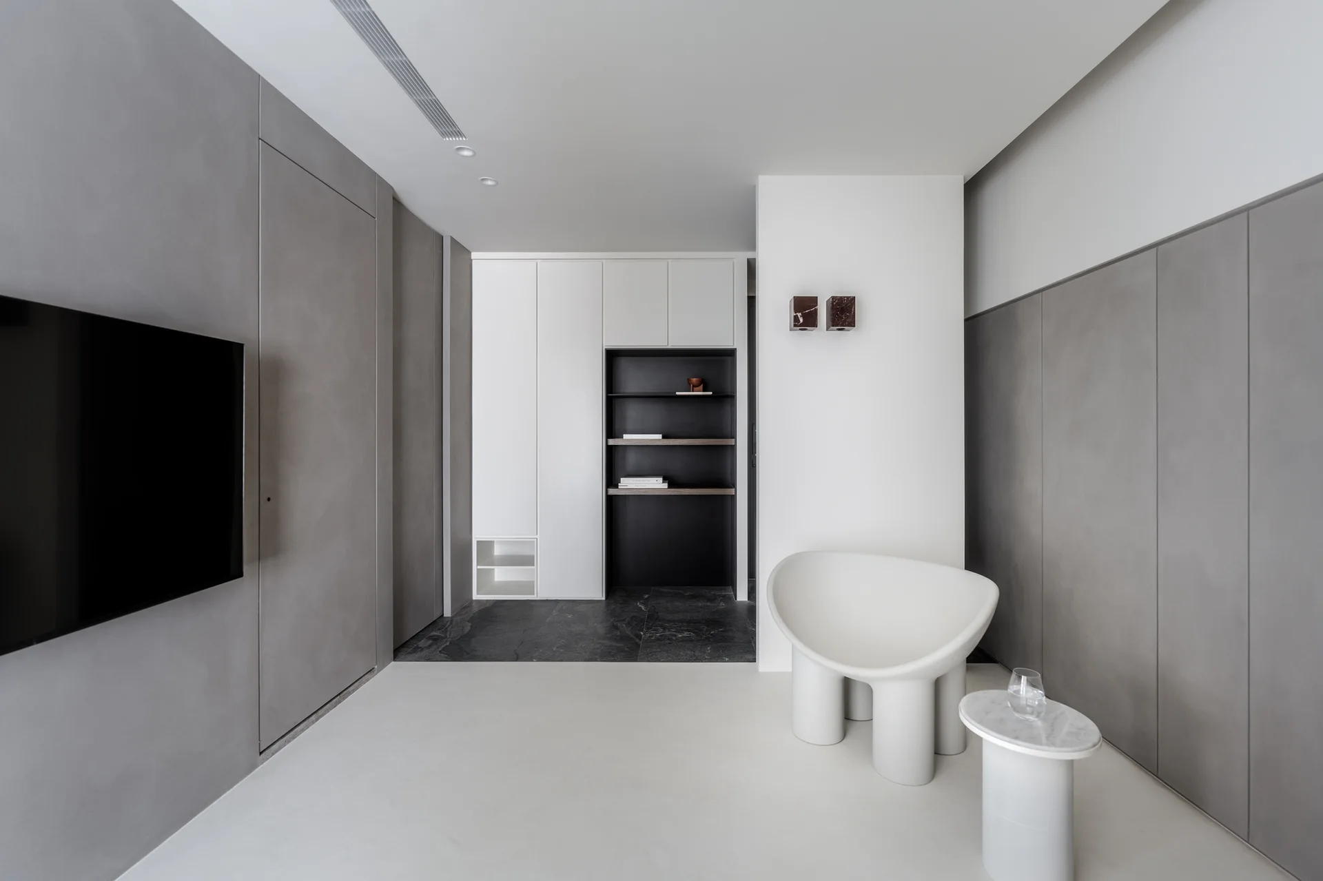 A minimalist room featuring a white sculptural armchair, a marble side table, smooth grey wall panels, and built-in white and black cabinetry.