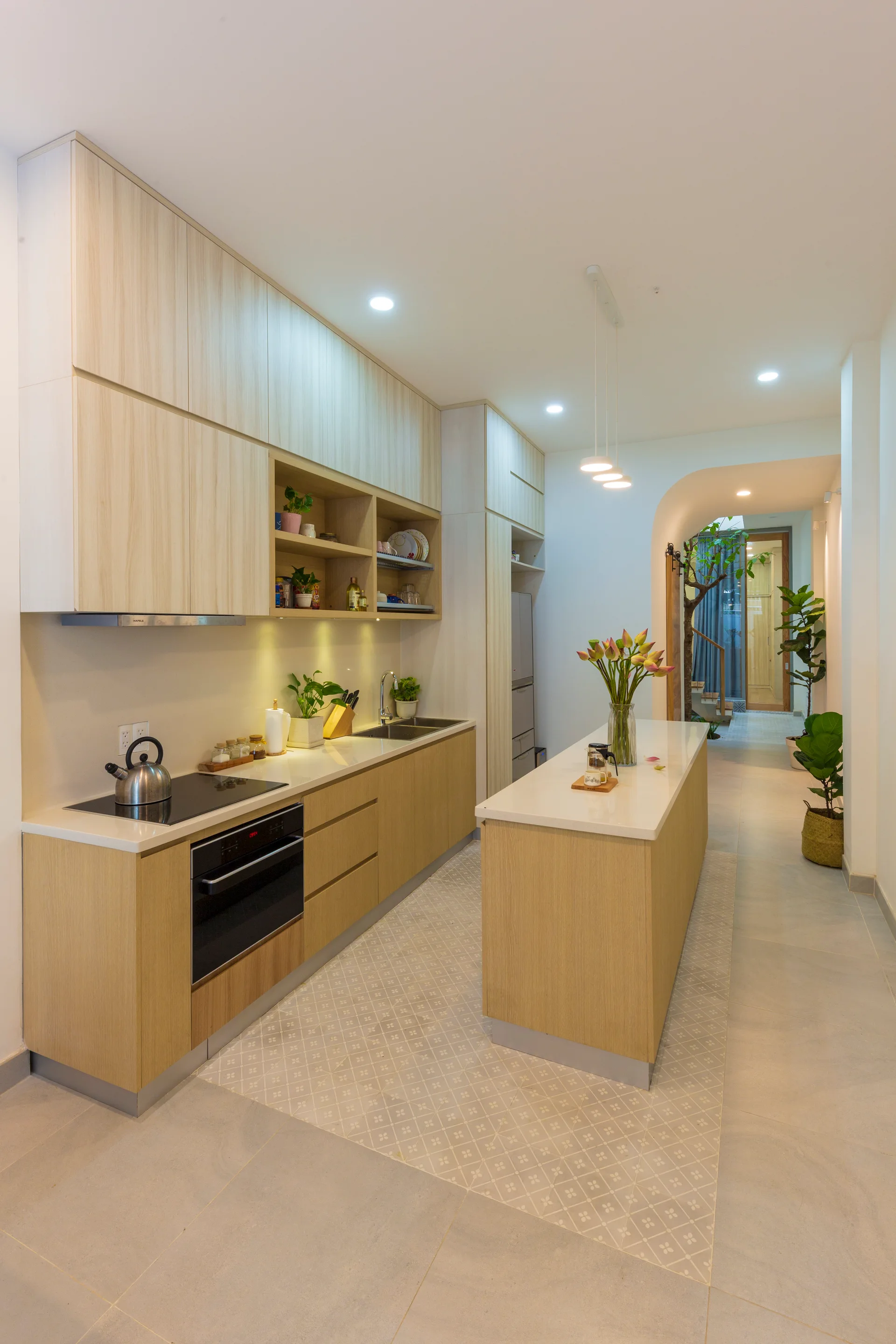 A long contemporary kitchen featuring light wood flat-front cabinets, white countertops, a central island, and a patterned tile floor inlay.