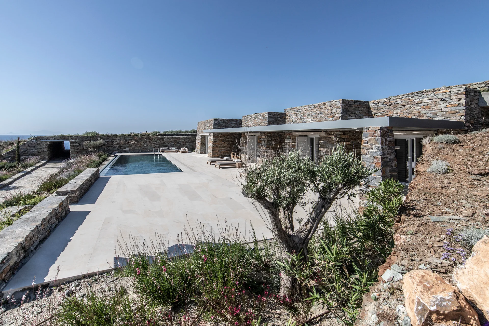 A rectangular swimming pool sits on a large beige stone patio next to a modern stone-walled house with a flat metal canopy under a clear blue sky.