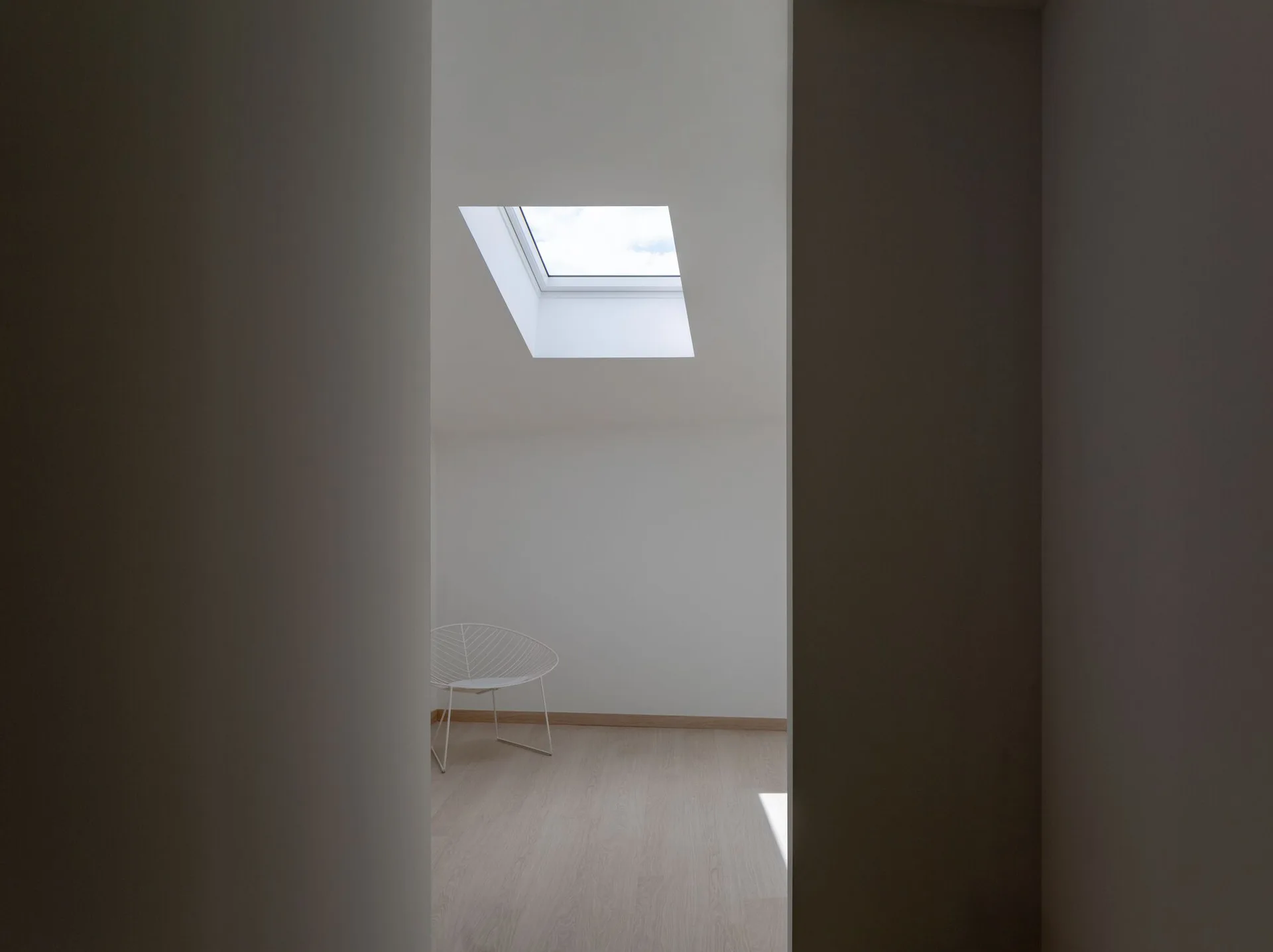 A view through a dark doorway into a minimalist white attic room featuring a skylight and a single white wire chair on light wood floors.