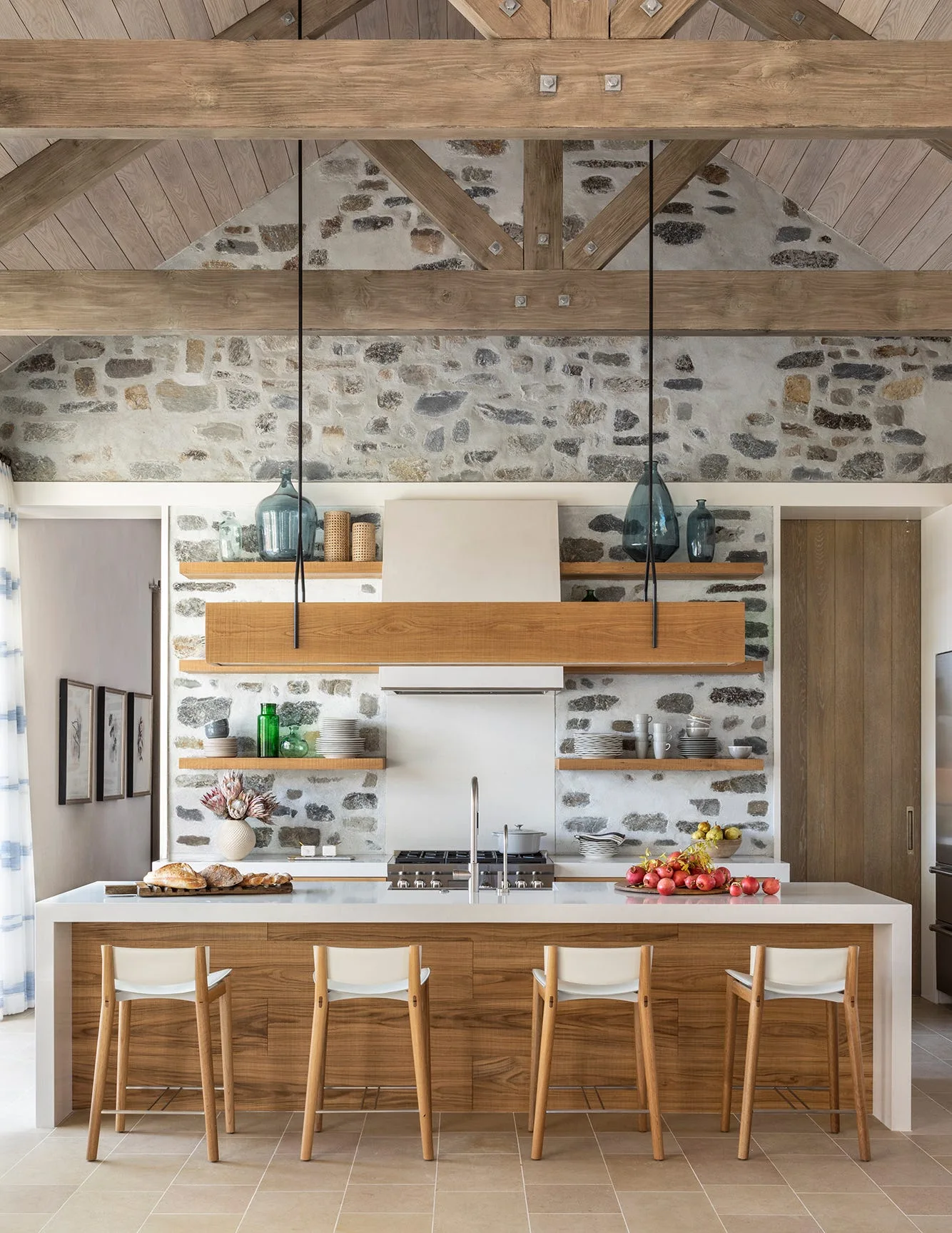 A rustic kitchen features a wood-clad island with white stools, floating wooden shelves against an exposed stone wall, and a vaulted timber ceiling.