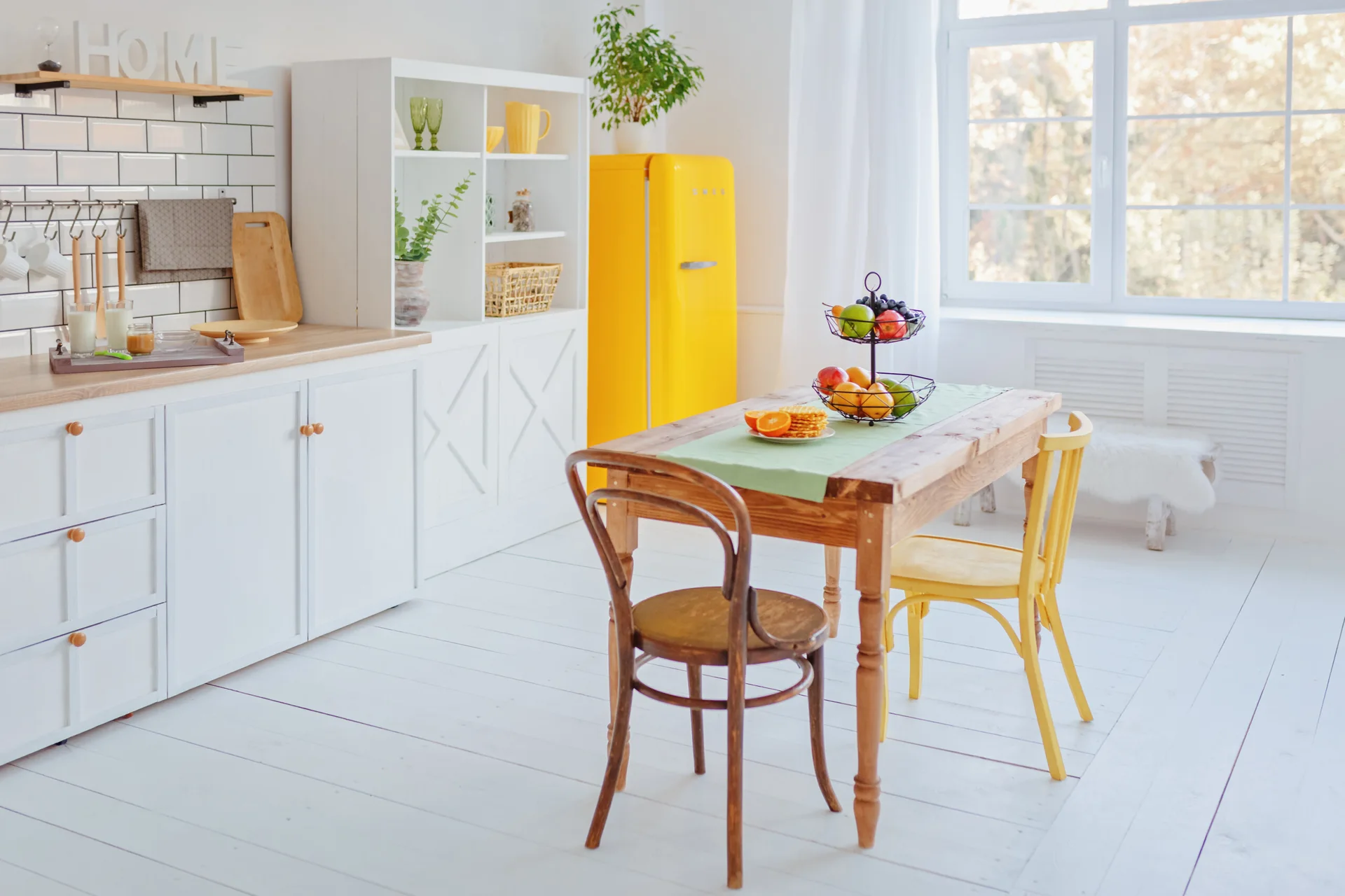 A bright white kitchen featuring a retro yellow refrigerator, a wooden dining table with mismatched chairs, and white subway tile backsplash.