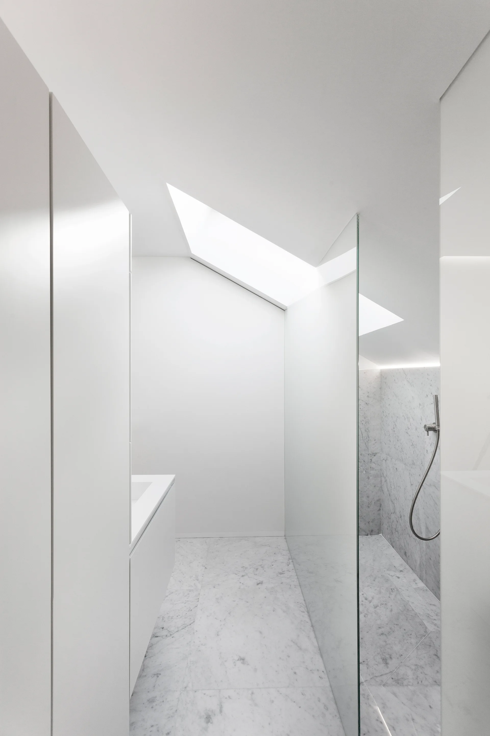 A minimalist white bathroom featuring a walk-in shower with a glass partition, veined marble flooring, and a large skylight in the sloped ceiling.