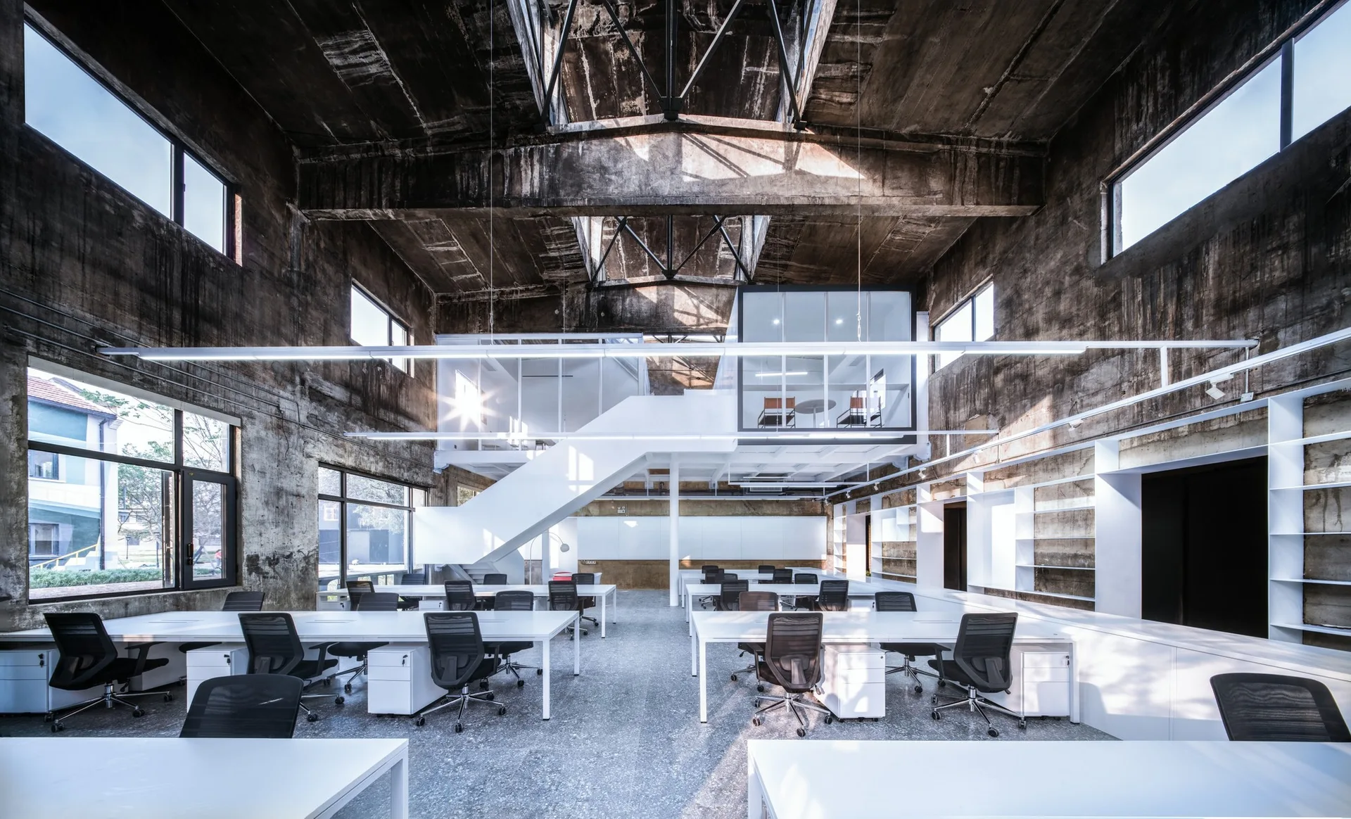 An industrial open-plan office features rows of white desks and black chairs under a raw concrete ceiling and a modern white mezzanine.
