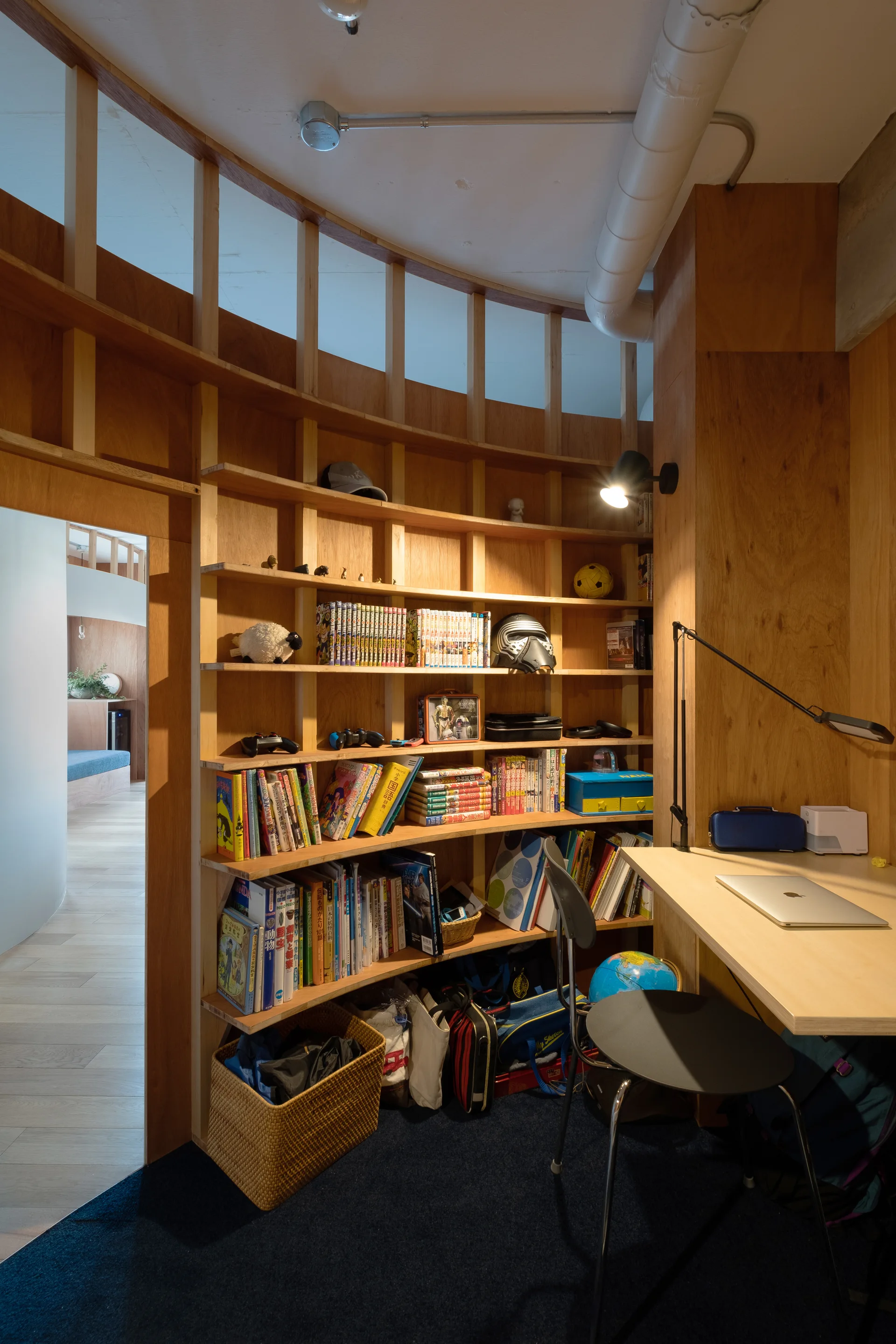 A home office features a curved wooden built-in bookshelf filled with books, a small desk with a laptop, and warm wood wall paneling.