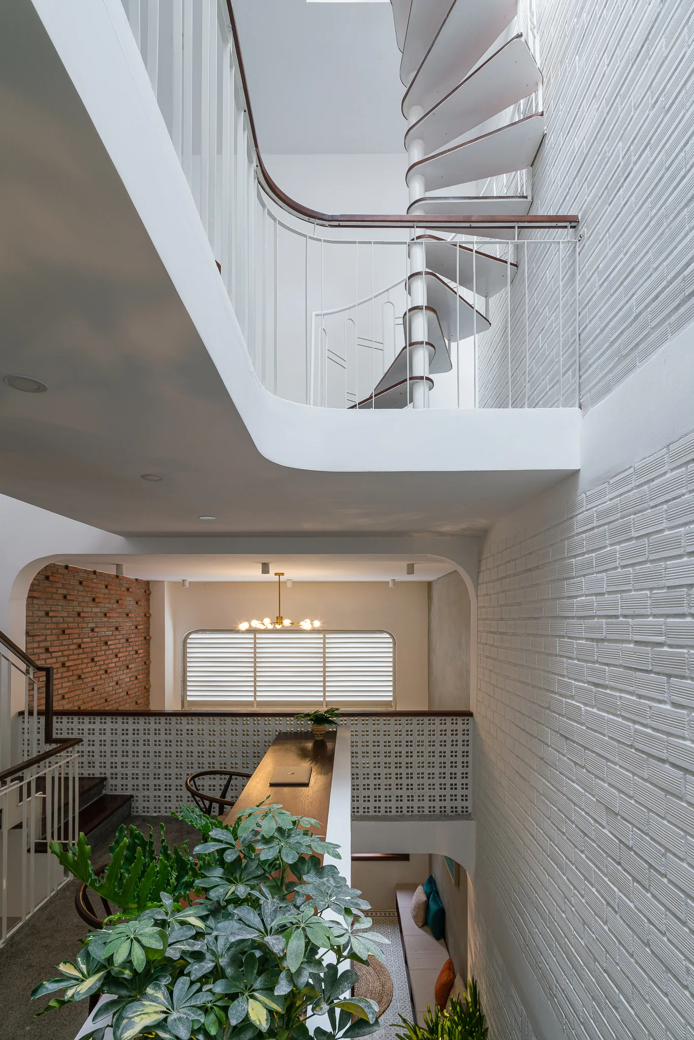 A multi-level interior features a white spiral staircase on the upper landing, a long wooden desk on the mezzanine, and a patterned breeze block partition.