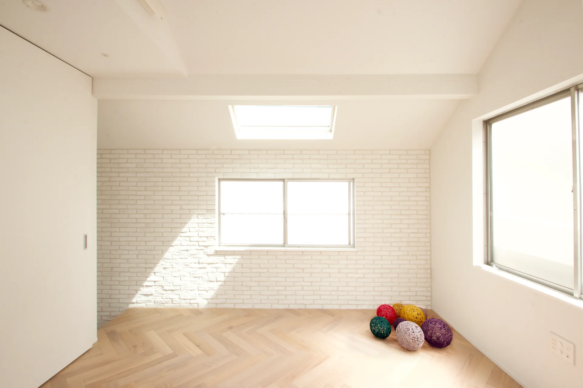 An empty minimalist room with a white brick wall, ceiling skylight, and light-toned wood flooring laid in a herringbone pattern.