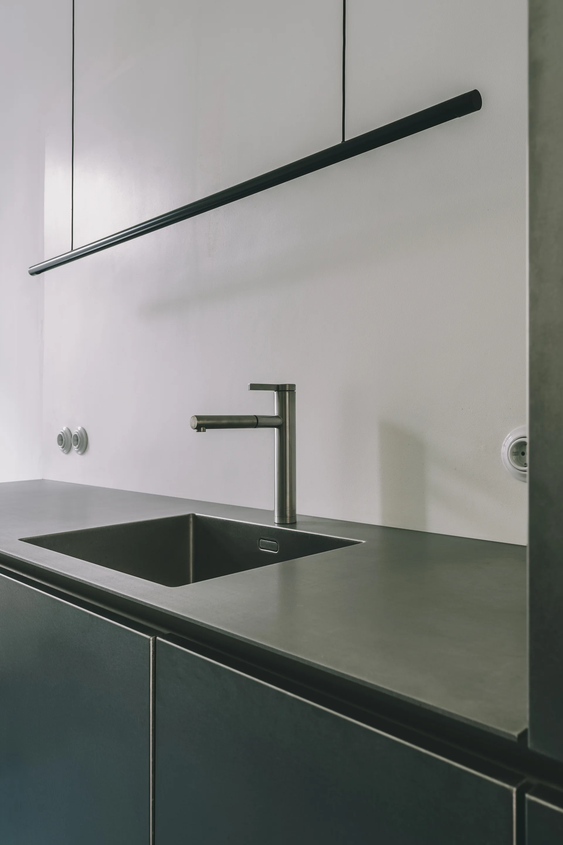 A close-up of a minimalist kitchen with a grey countertop, integrated sink, brushed metal faucet, and dark matte cabinetry.